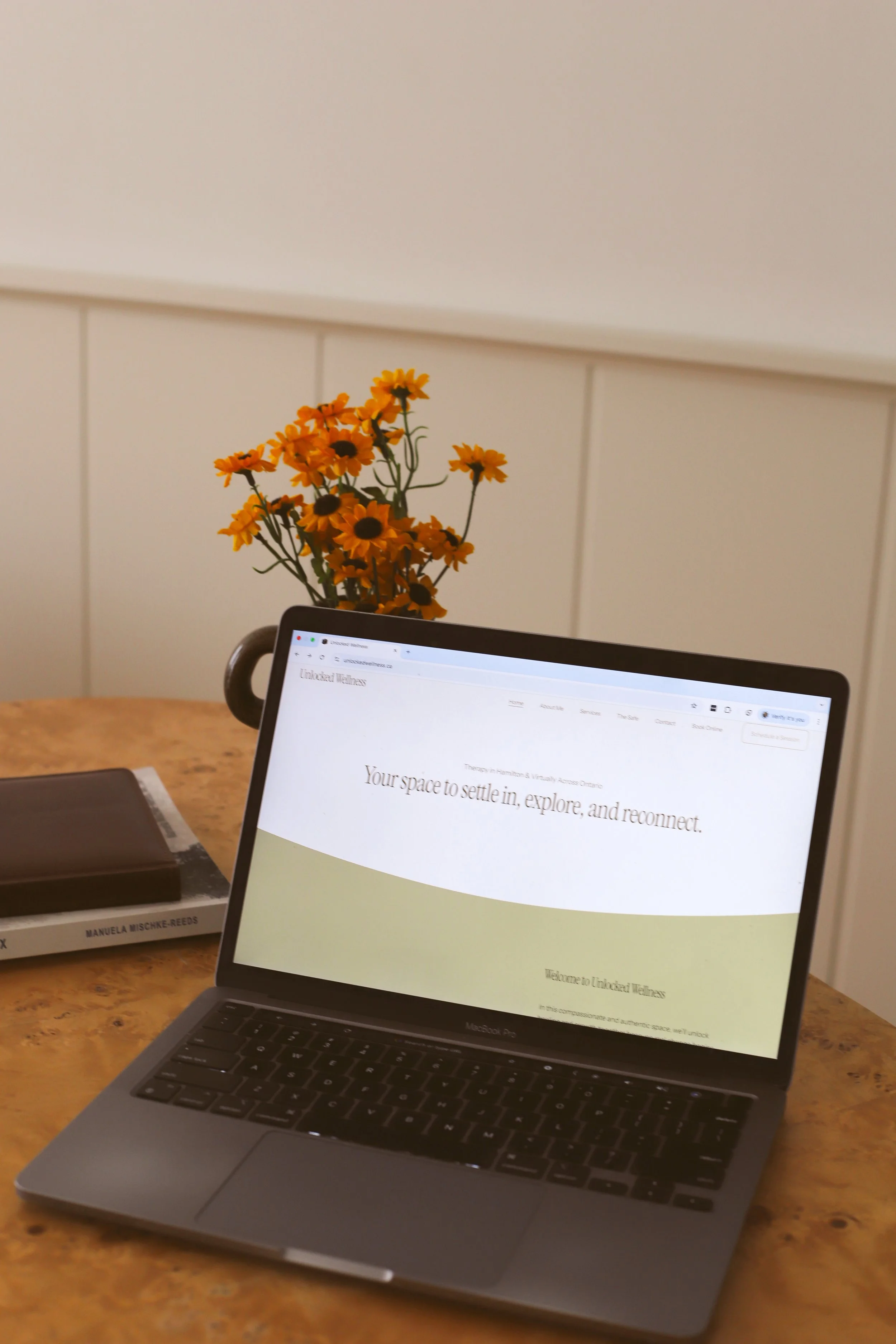 A wooden table with an open MacBook Pro displaying a website, a closed book, and a small vase with yellow flowers.
