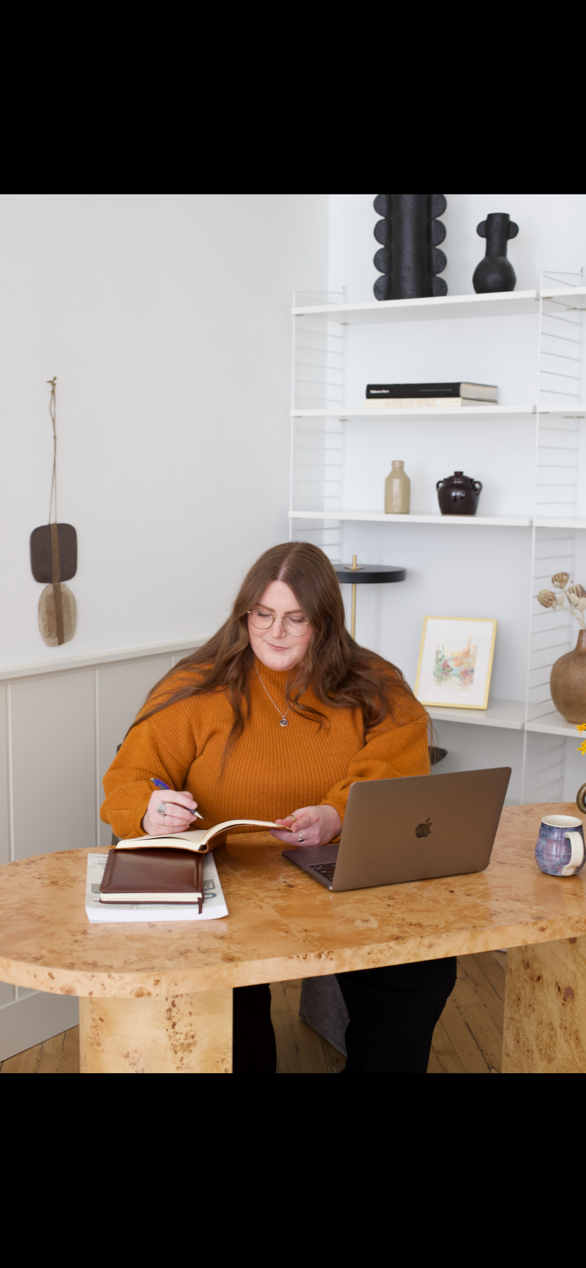 A woman with long brown hair and glasses sitting at a wooden desk, writing in a notebook with a pen, with a laptop, mug, and closed book on the desk. Behind her is a white shelf with decorative items, a small painting, and a vase.