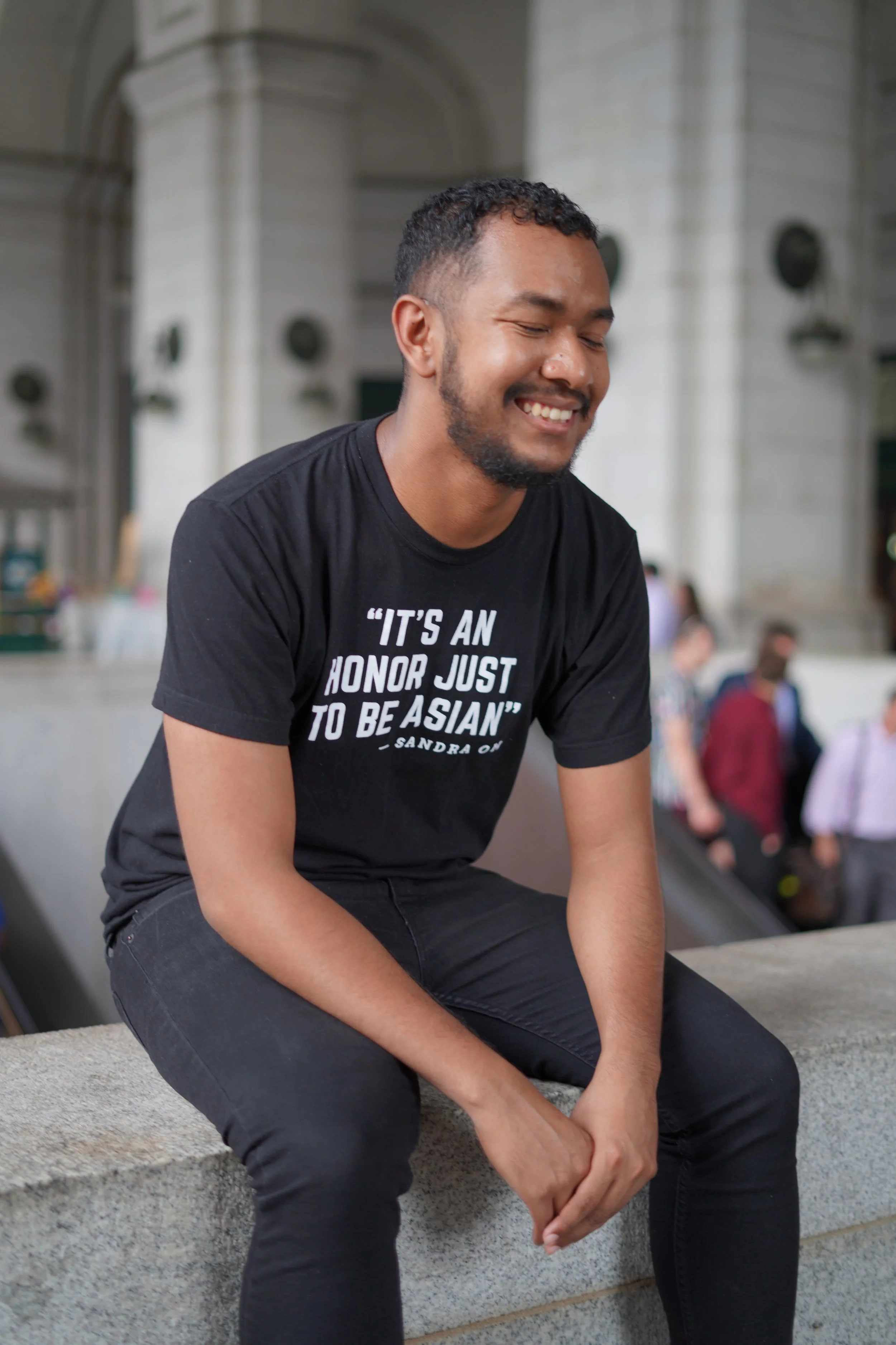 A young man sitting outdoors on a stone ledge, smiling with eyes closed, wearing a black T-shirt with white text and black pants, with a blurred background of other people and architecture.