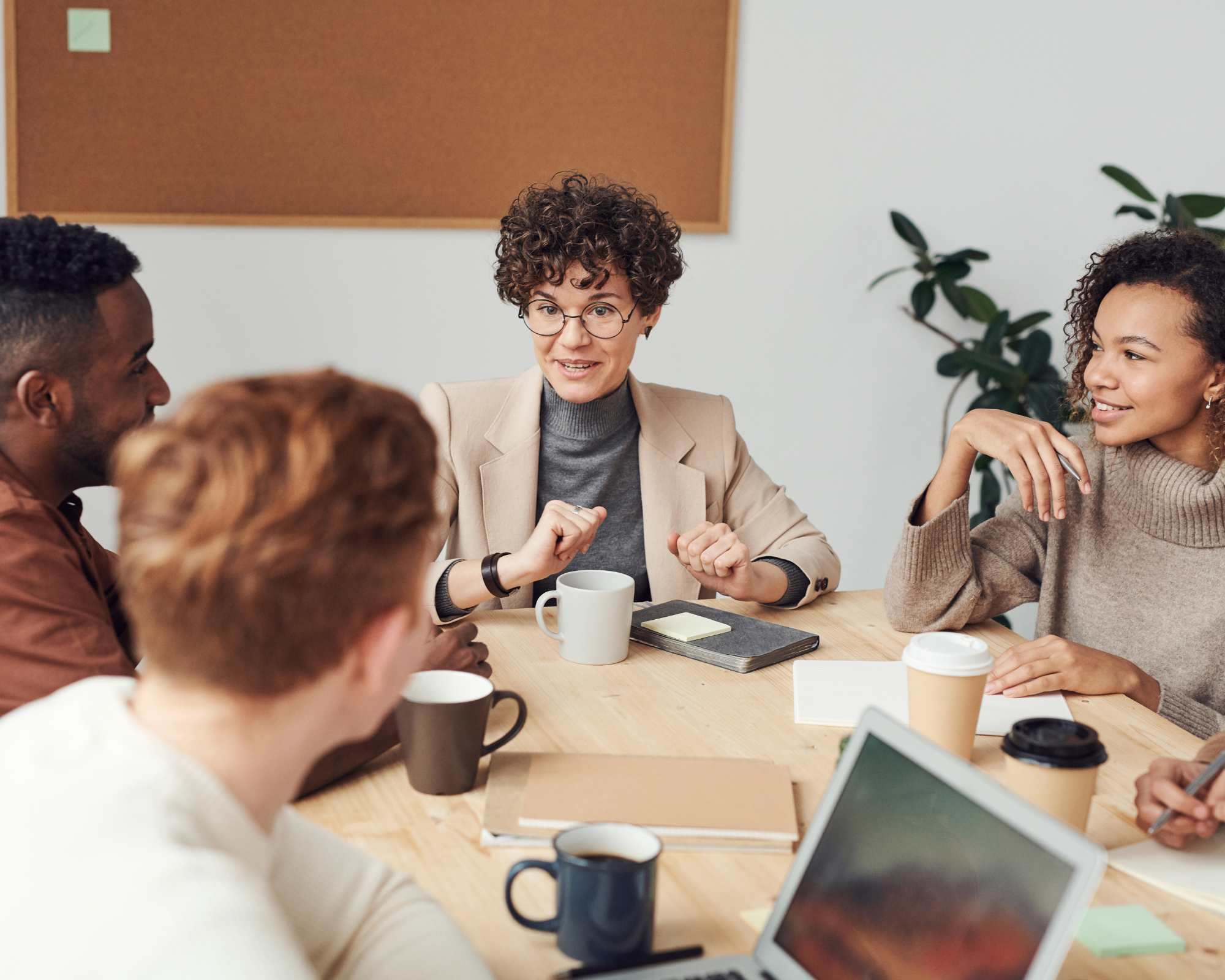 A diverse group of five people engaged in a lively discussion in a meeting room, with notes, cups, and laptops on the table.