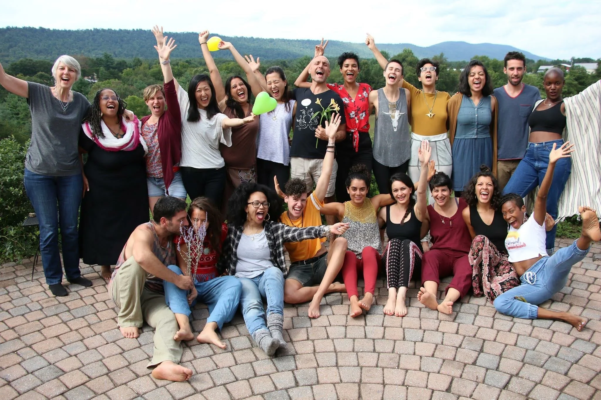 A large group of diverse young adults posing together outdoors on a paved area with trees and mountains in the background, laughing and cheering, some sitting on the ground and others standing, many raising their hands or holding balloons and flowers.