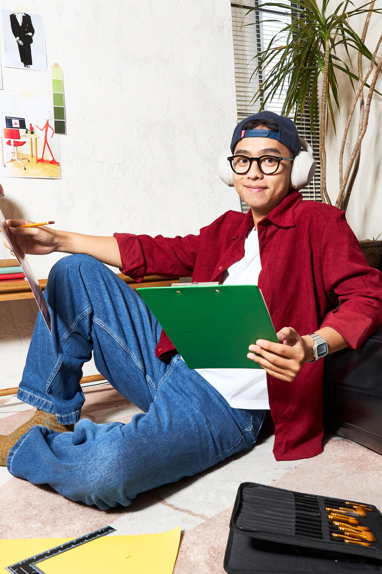 Young man sitting on the floor with a green clipboard and headphones, surrounded by drawing tools and papers, in a room with a textured white wall, a tall plant, and window blinds.