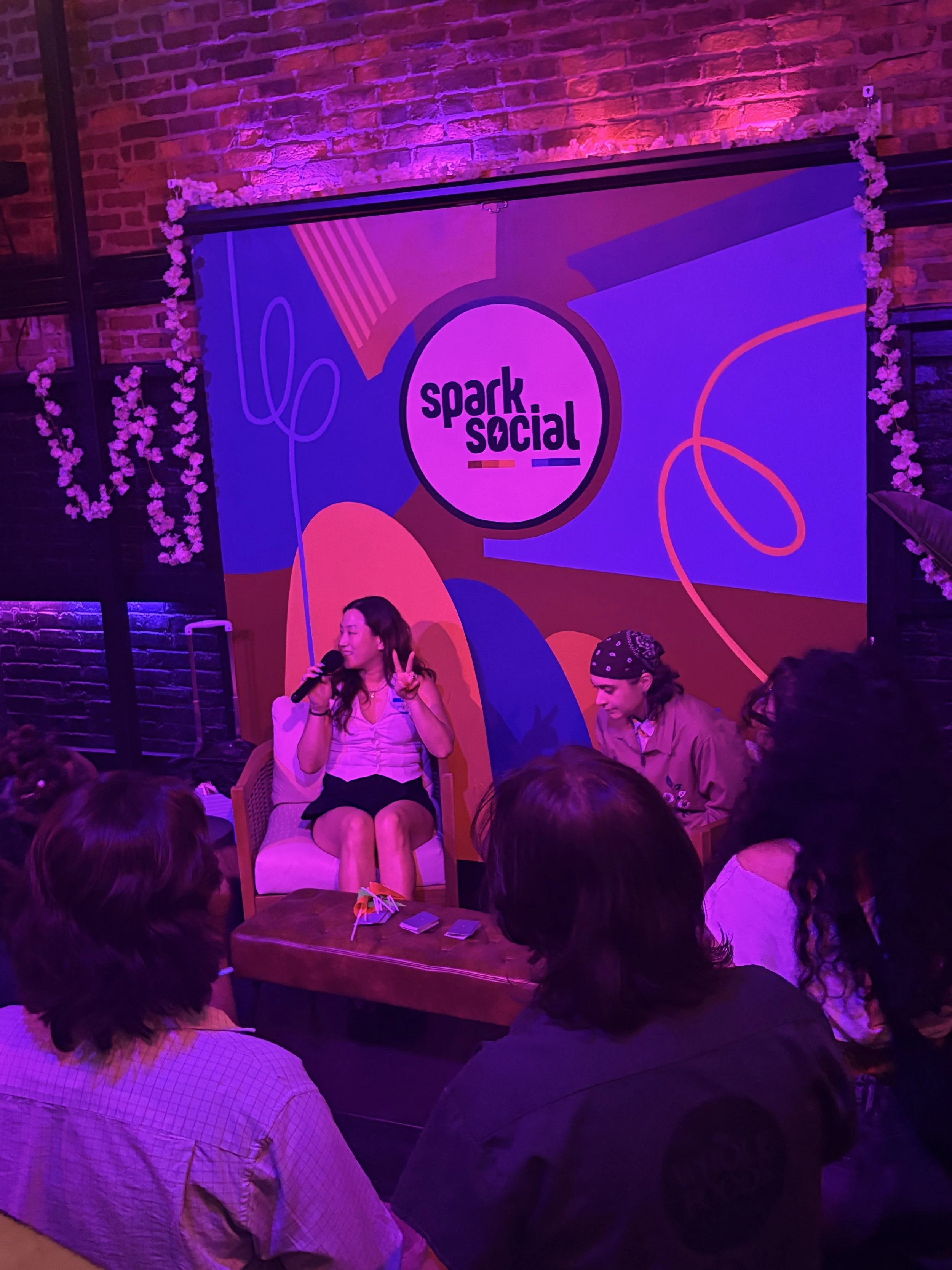 A woman sitting on a chair holding a microphone and making a peace sign during a panel or discussion at an event called Spark Social, with a colorful vibrant backdrop and an audience in front.