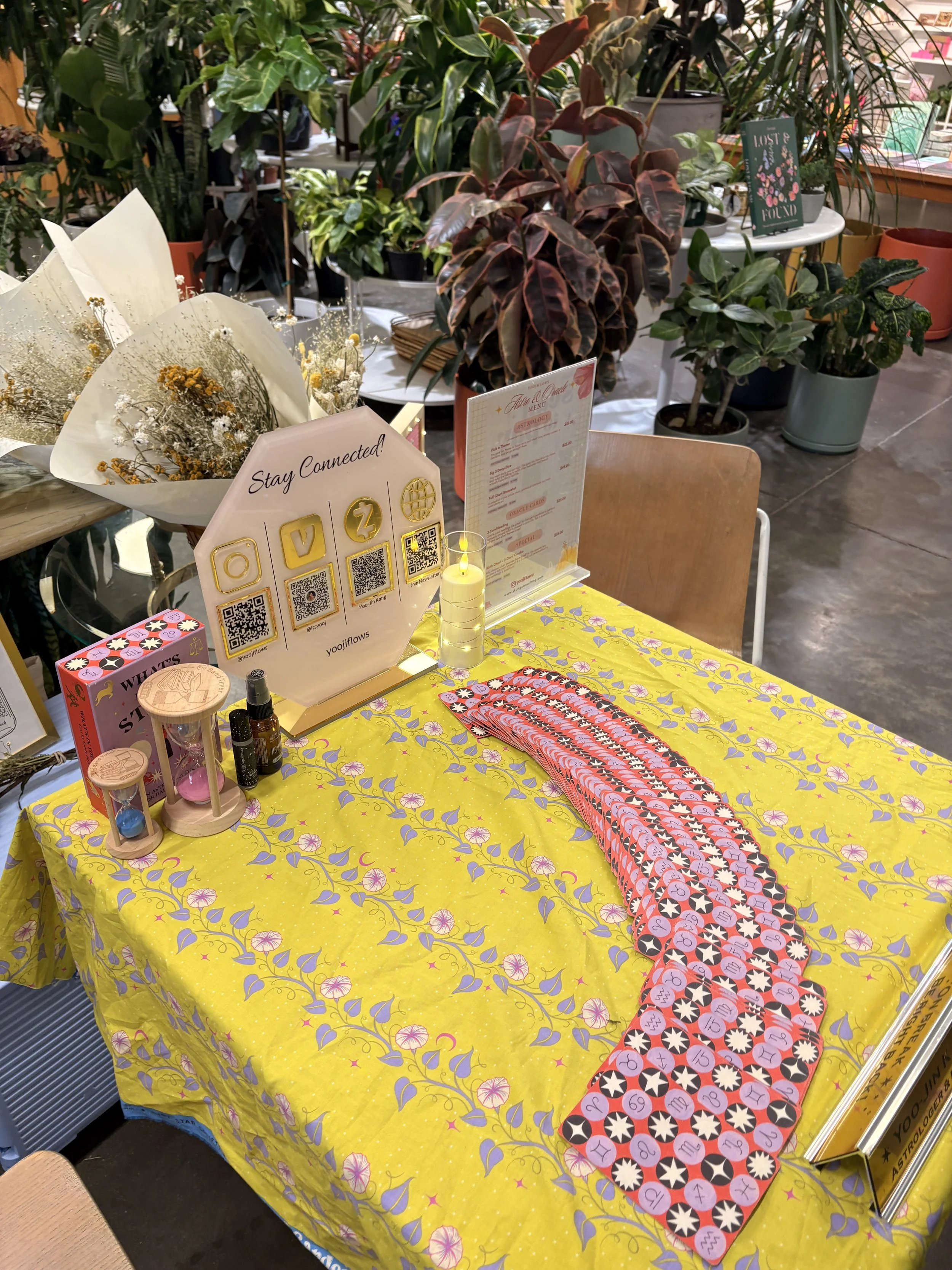 A table covered with a yellow floral tablecloth, displaying lottery or bingo-like game cards with numbers and stars, along with various small decorative items, framed signs with QR codes, a lit candle, and floral arrangements, set in a setting with a lush background of potted plants and greenery.