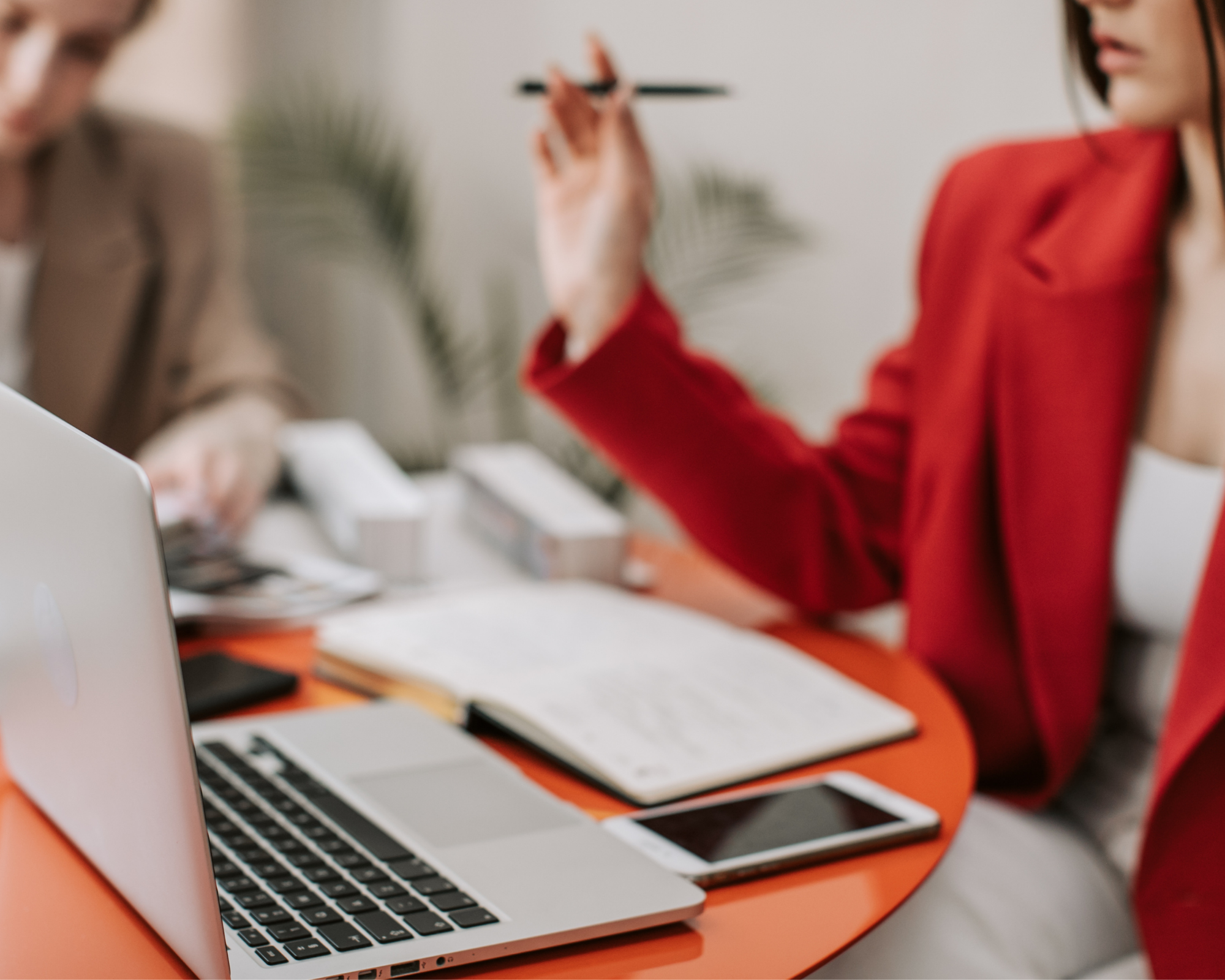 Business meeting with a woman in a red blazer gesturing while a man in a beige blazer is seated nearby at a table with a laptop, smartphone, notebook, and various office supplies.