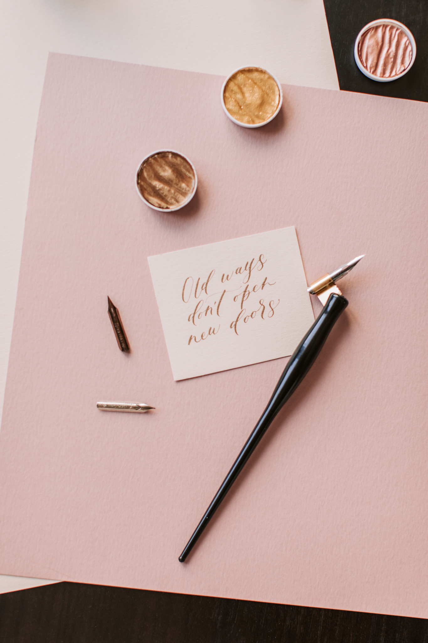A desk with pink and black papers, a calligraphy pen, small containers of metallic paint, and a handwritten note that says 'Old ways don't open new doors'.