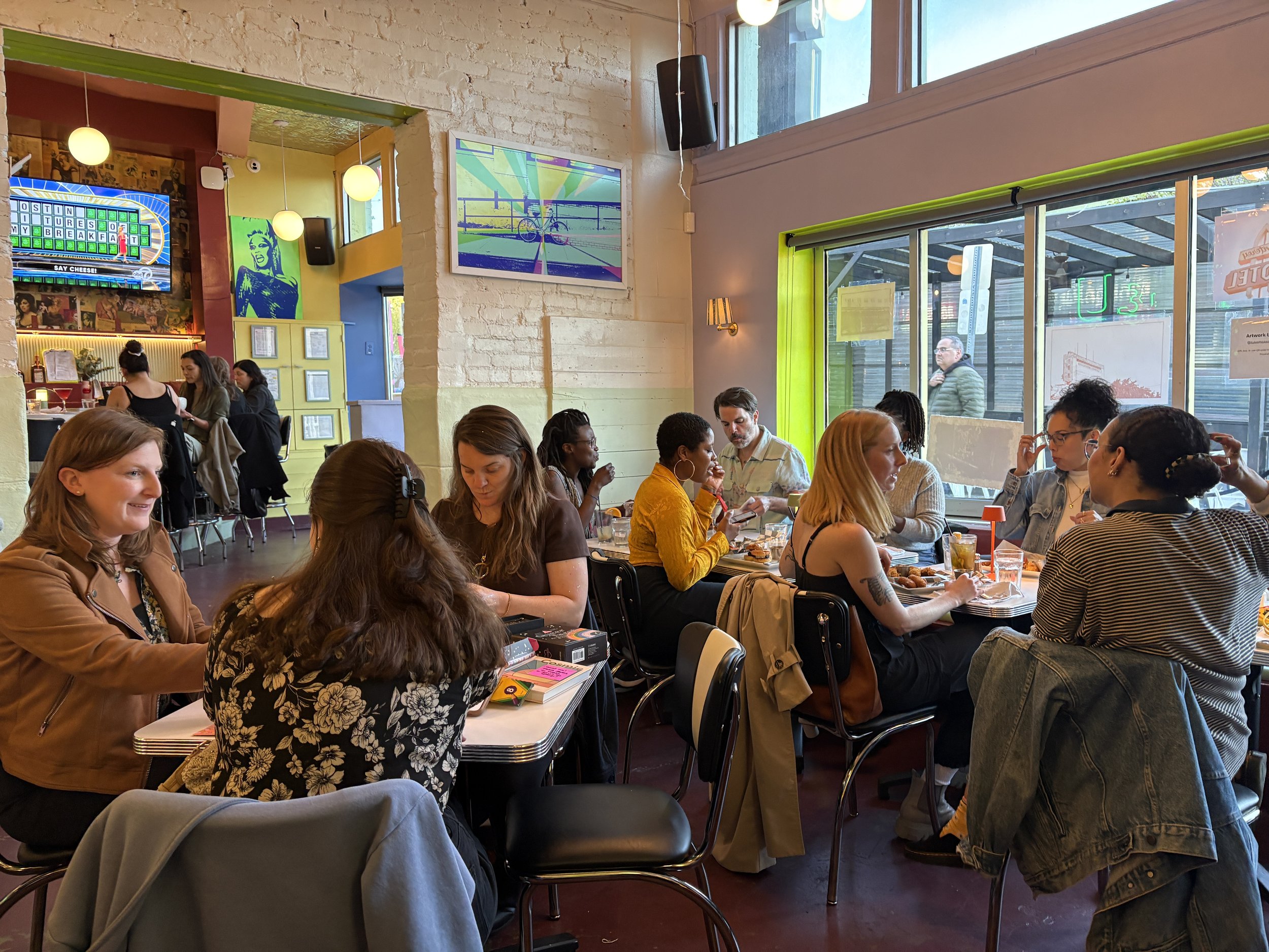 People sitting at tables inside a casual restaurant or cafe, socializing and dining. Bright decor with colorful artwork and large windows letting in natural light.