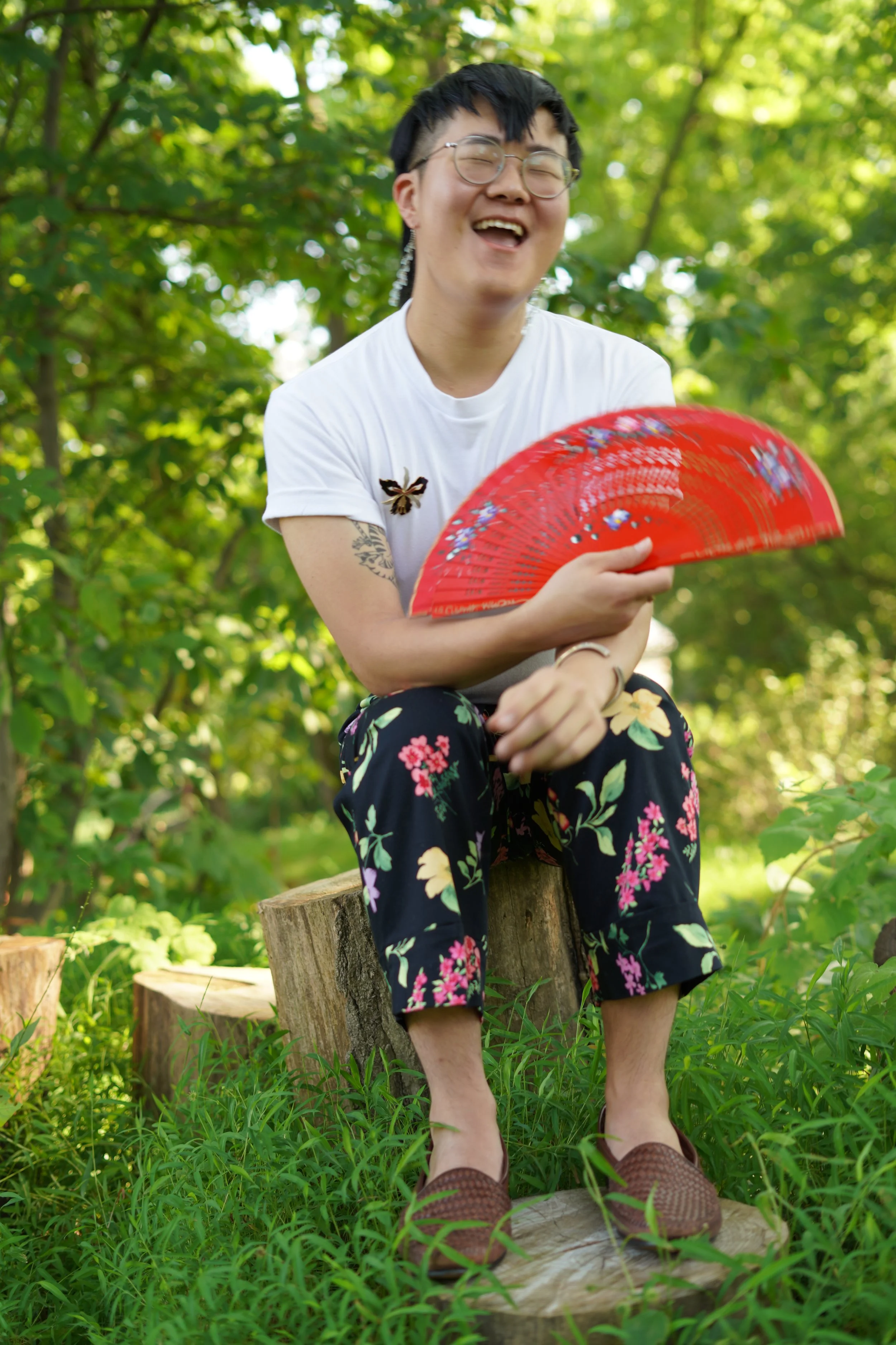 A person sitting outdoors on a tree stump, holding a red folding fan, smiling and laughing, with trees and green foliage in the background.