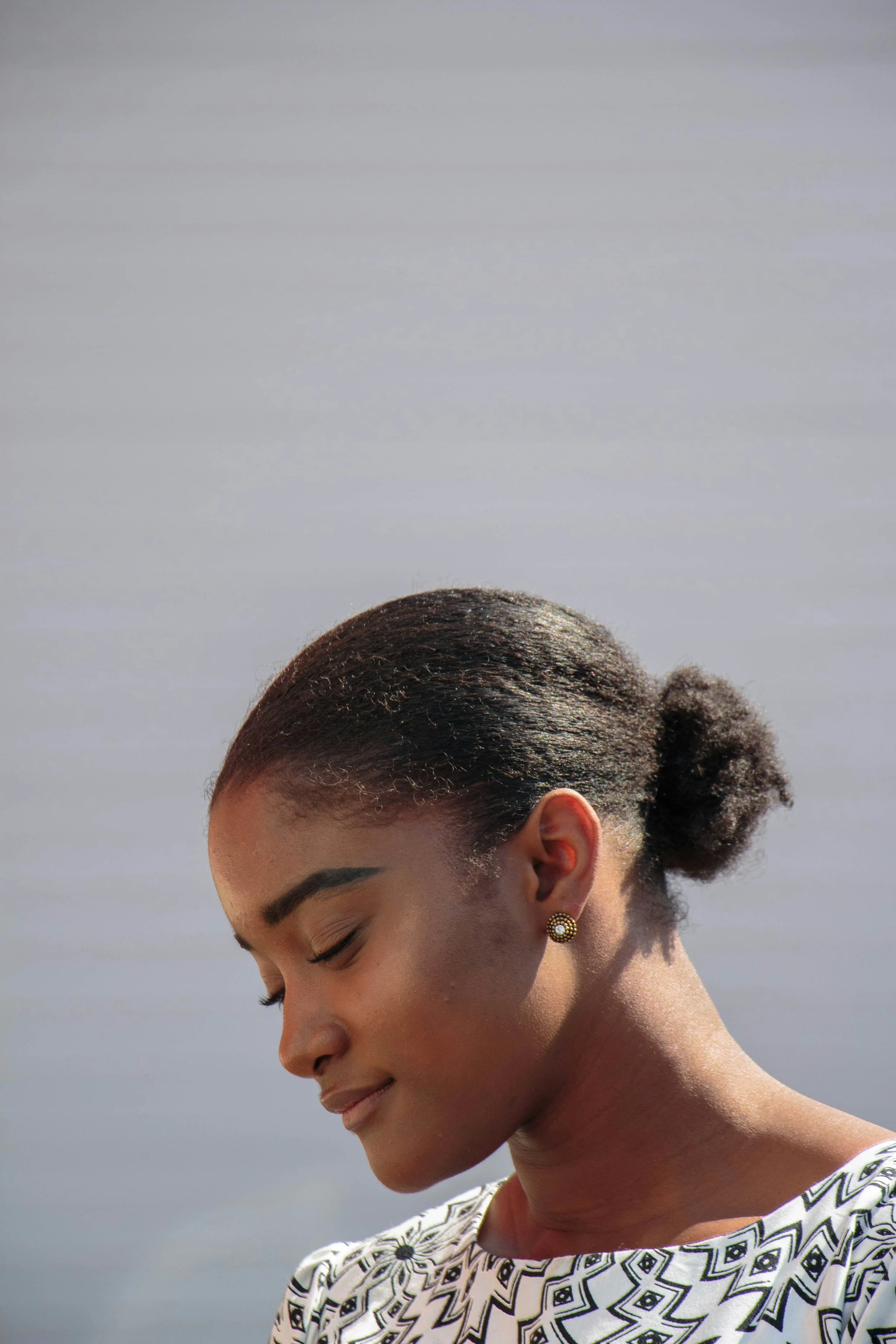 Profile of a woman with closed eyes, dark hair in a bun, wearing patterned top and gold earrings, outdoors near water.