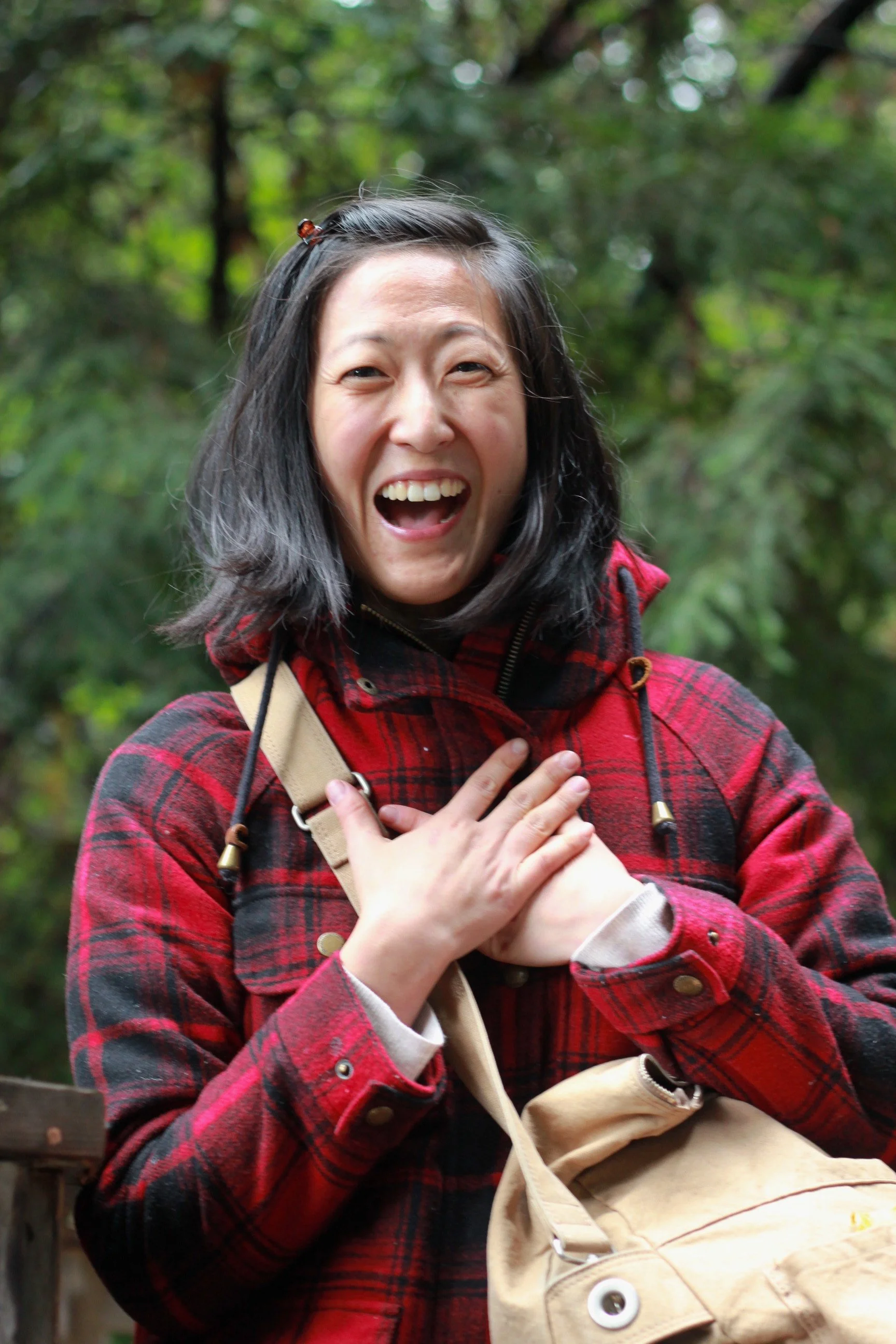 A woman in a red plaid jacket with a beige shoulder bag, smiling excitedly outdoors with a green, tree-filled background.