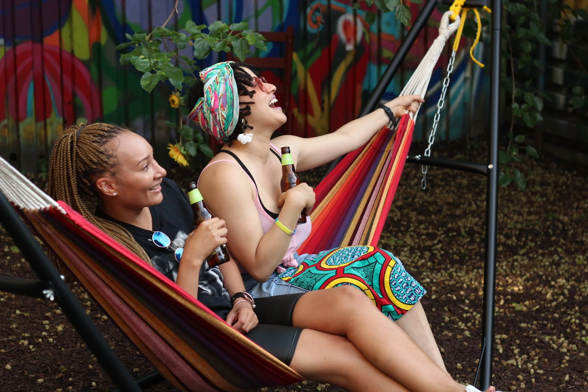 Two women relaxing on a swing in a colorful outdoor setting, holding beer bottles, laughing, and enjoying each other's company.
