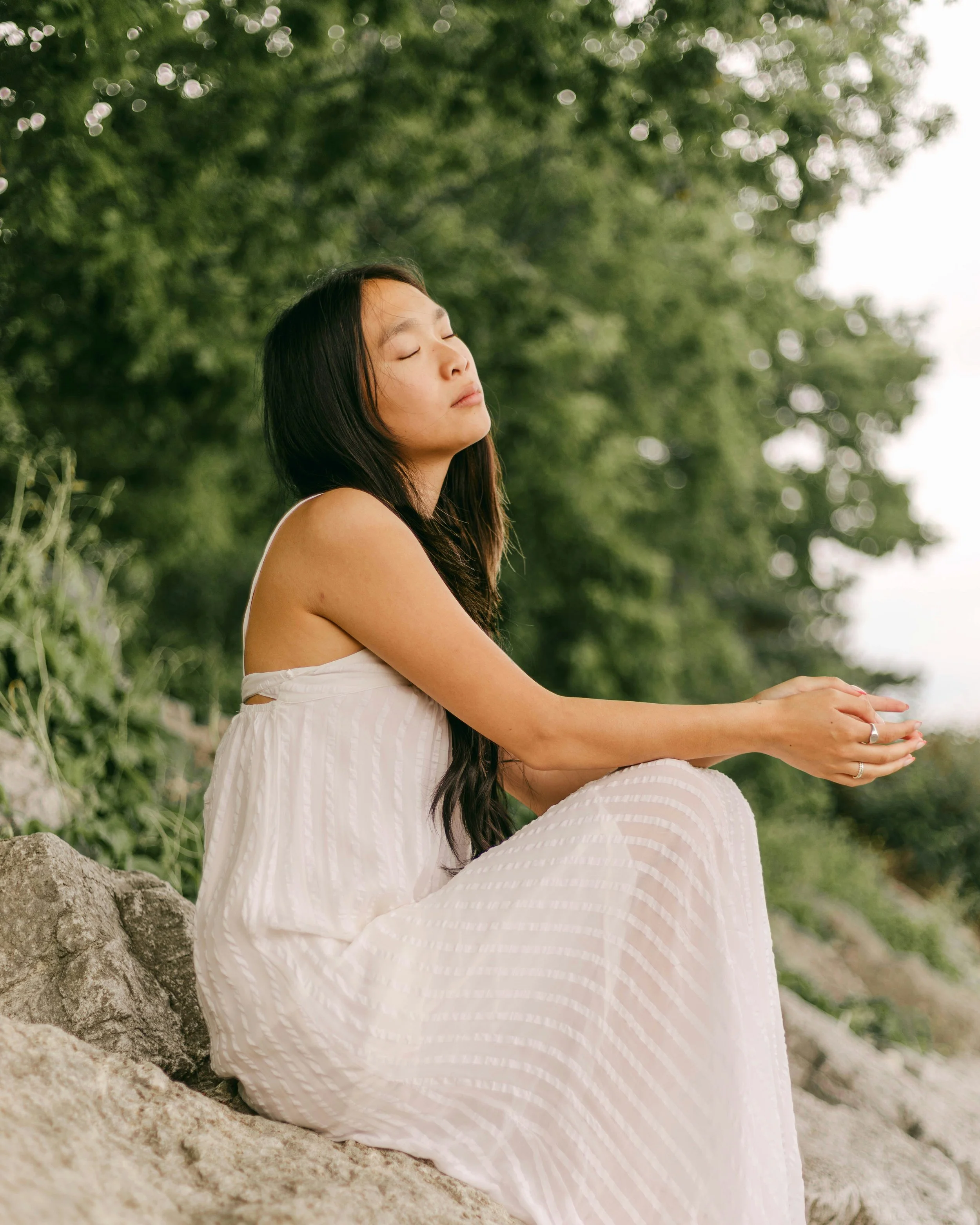 A woman with long dark hair, wearing a white dress, sits outdoors with eyes closed and hands resting on knees, surrounded by greenery.