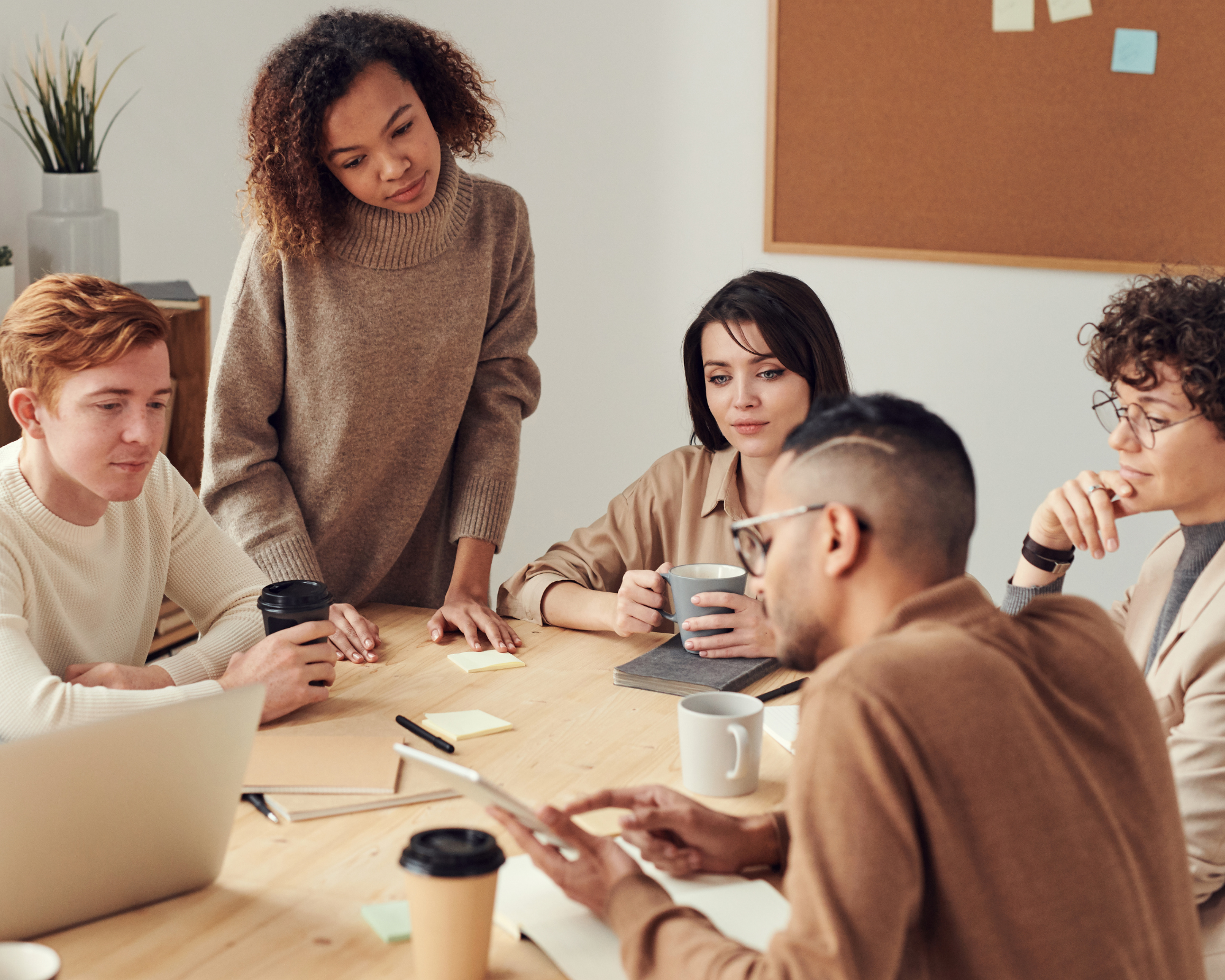 A diverse group of young adults gathered around a wooden table in a casual meeting or study session. They are engaged in discussion, with some looking at documents or notebooks, one person holding a coffee mug, and others listening attentively. The background features a minimalist decor with a cork bulletin board and a potted plant.