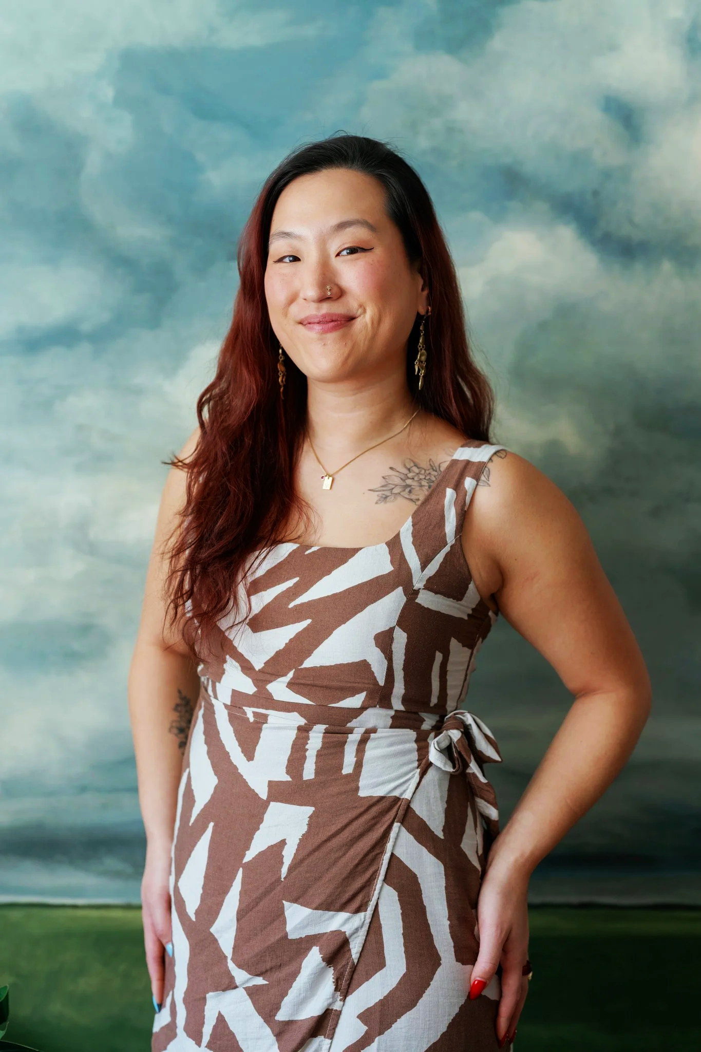 A woman with long wavy hair, wearing a beige and white patterned dress, standing in front of a cloudy sky background.