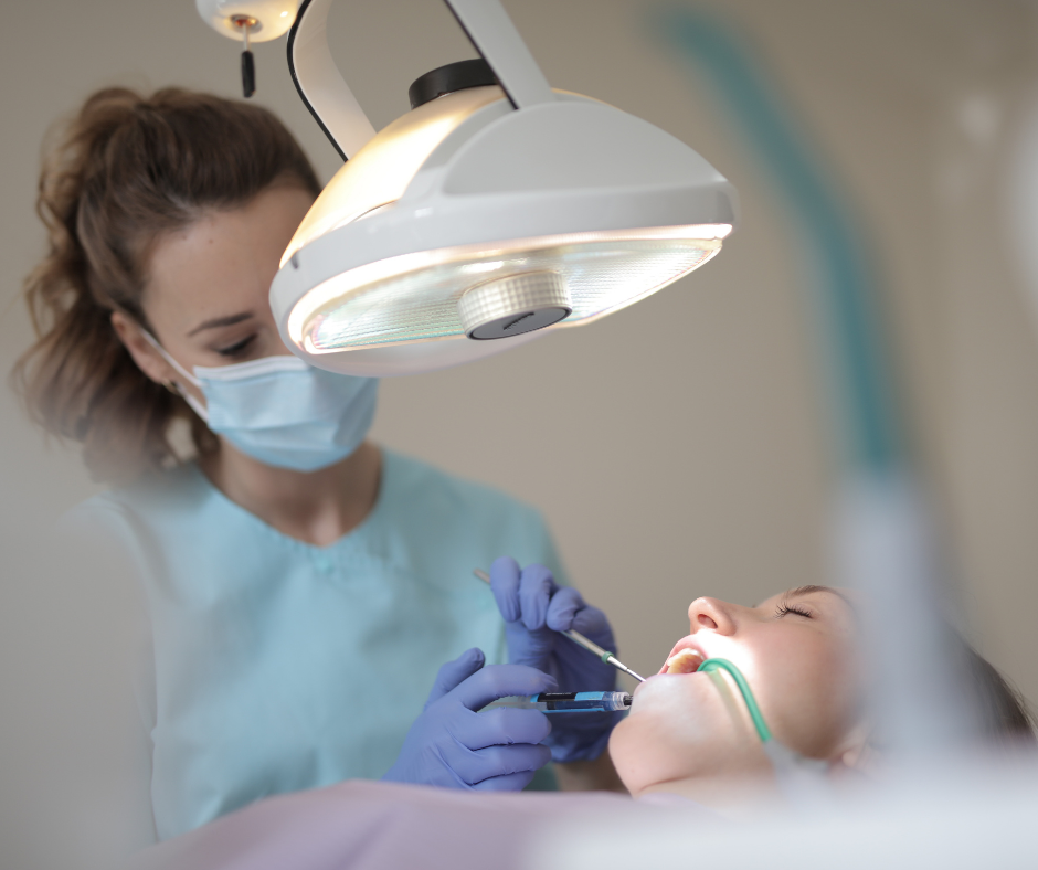 Dentist wearing gloves and a mask performing a dental procedure on a patient under an overhead exam light