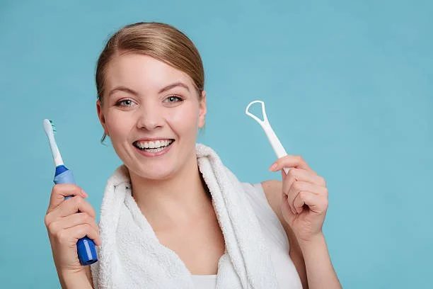 Smiling woman holding a tongue brush and electric toothbrush before cleaning her tongue.