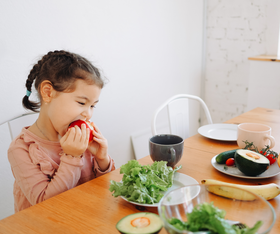 Young child eating a red apple at a kitchen table with healthy fruits and vegetables, illustrating nutrition’s role in children’s dental health