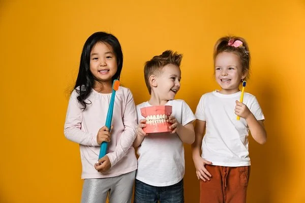 Three young children standing with dental props, including toothbrushes and a model of teeth, against a yellow background.