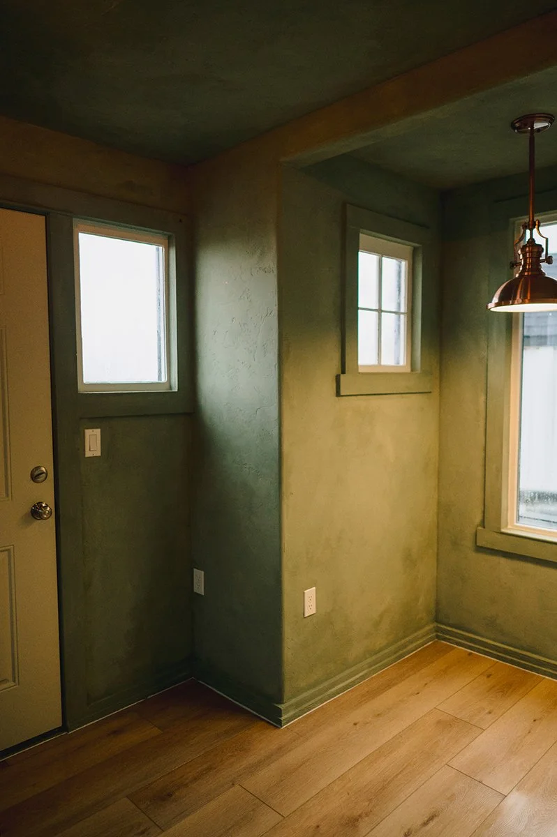 Vintage windows frame venetian textured walls finished with Color Atelier Cypress Ink limewash in 200, 150, and 100 percent layers, paired with Flooret Nakan flooring for a warm, functional artisan space.
