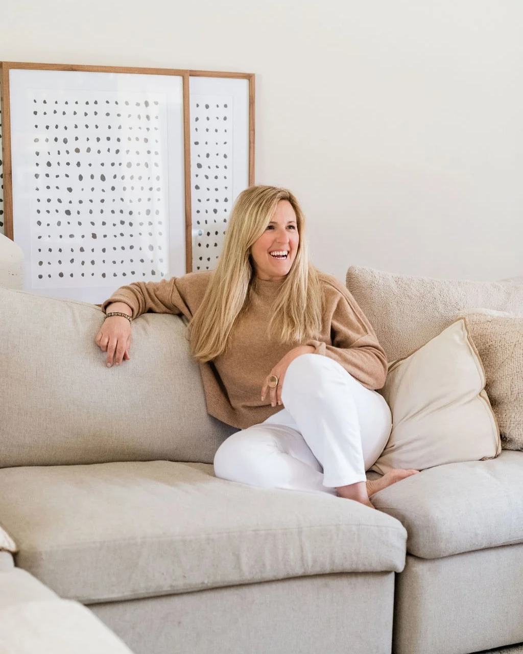 A woman with long blonde hair laughing while sitting on a beige couch in a living room.