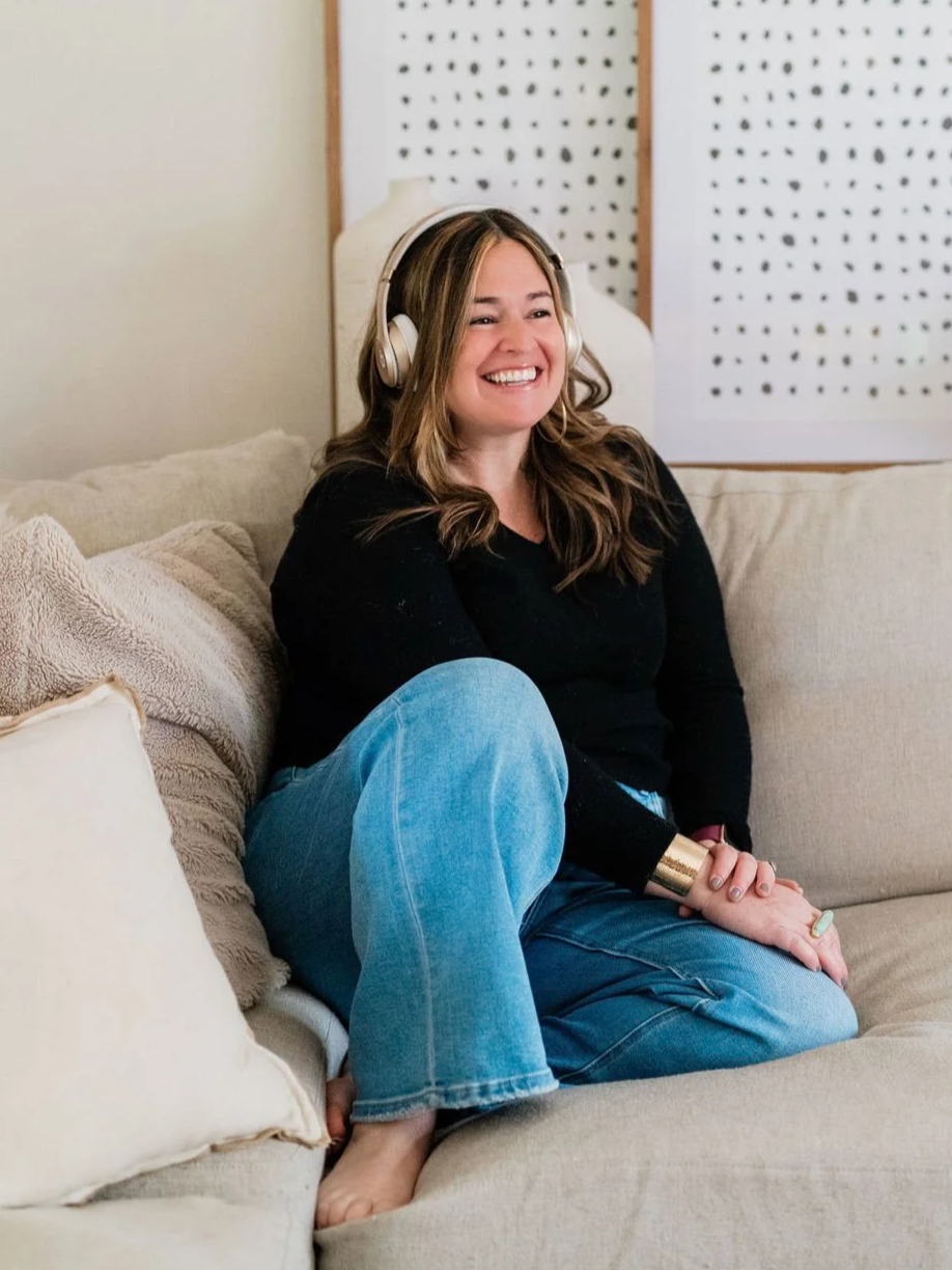 A woman with brown hair wearing headphones, a black sweater, and blue jeans, sitting on a beige sofa with pillows, smiling and enjoying music in a cozy room.