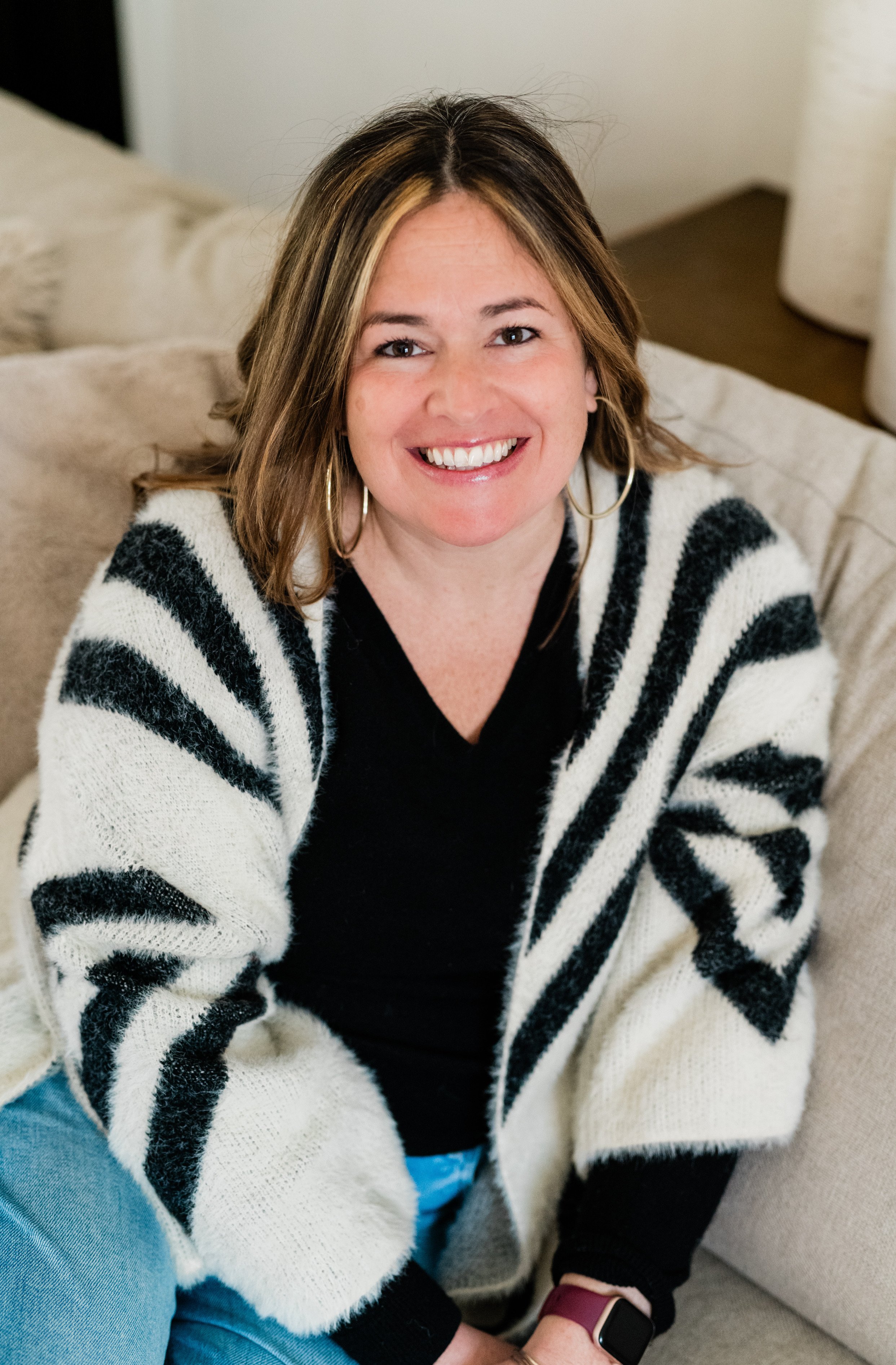 A woman with shoulder-length brown hair, wearing a black and white striped cardigan, black top, light blue jeans, and a smartwatch, smiling while sitting on a beige couch in a bright room.