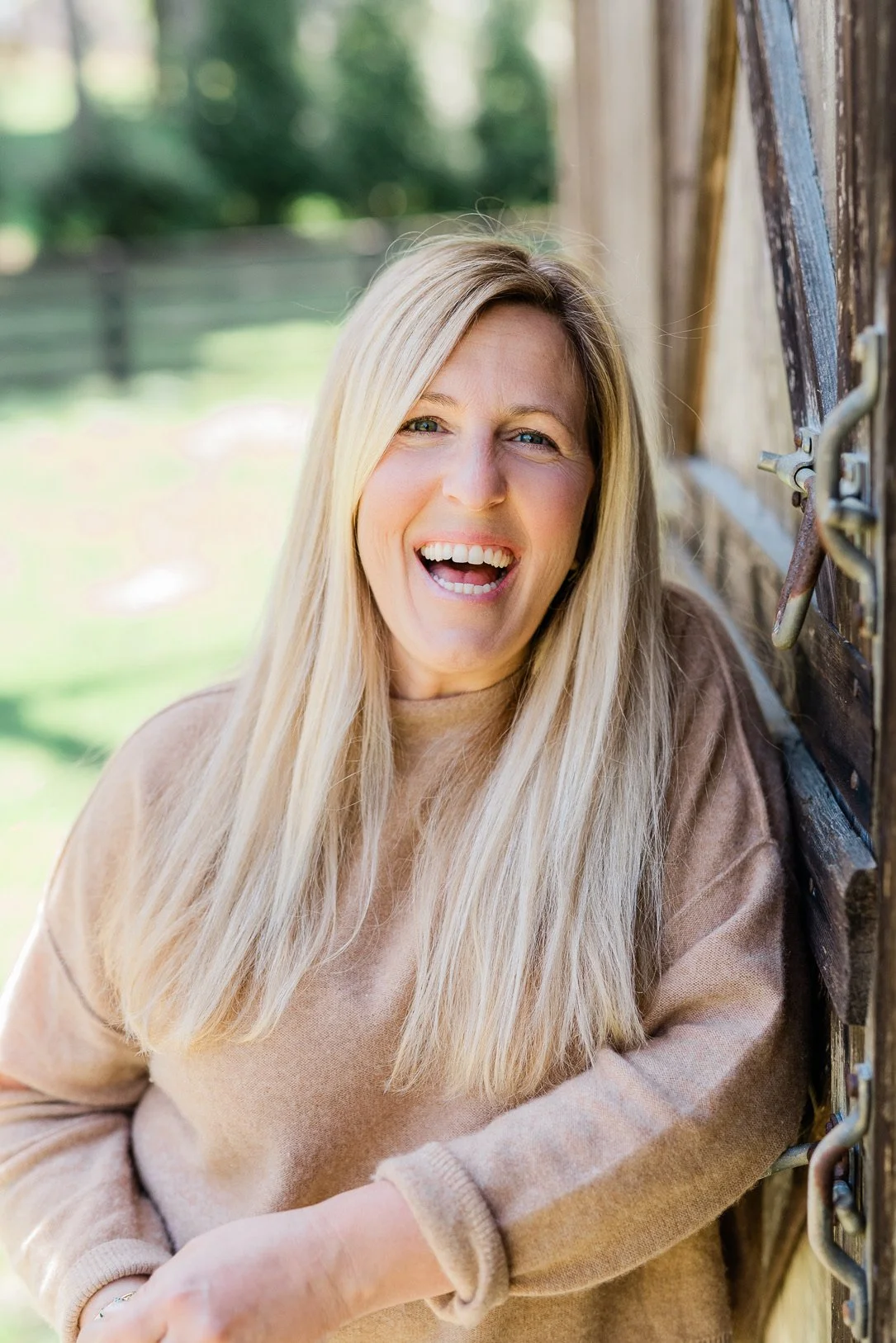A woman with long blonde hair smiling outdoors near a wooden barn.