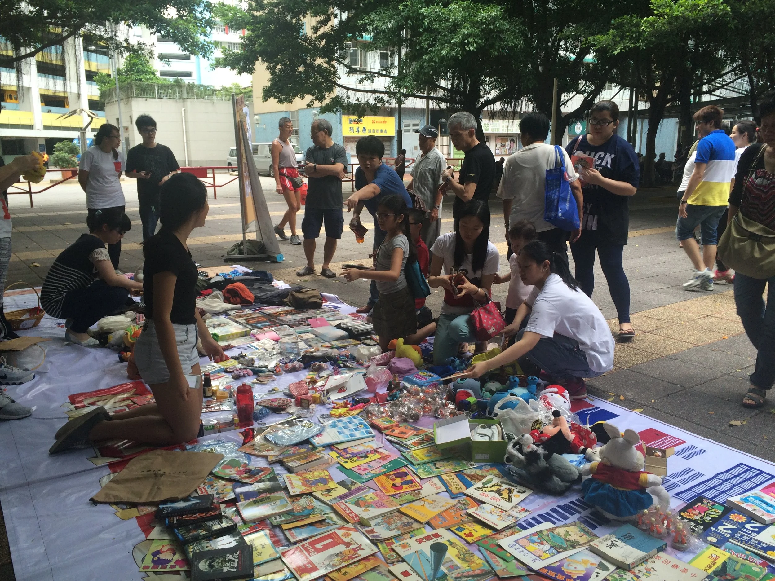 Outdoor flea market with people browsing toys, books, and clothes displayed on a large white sheet under a tree.