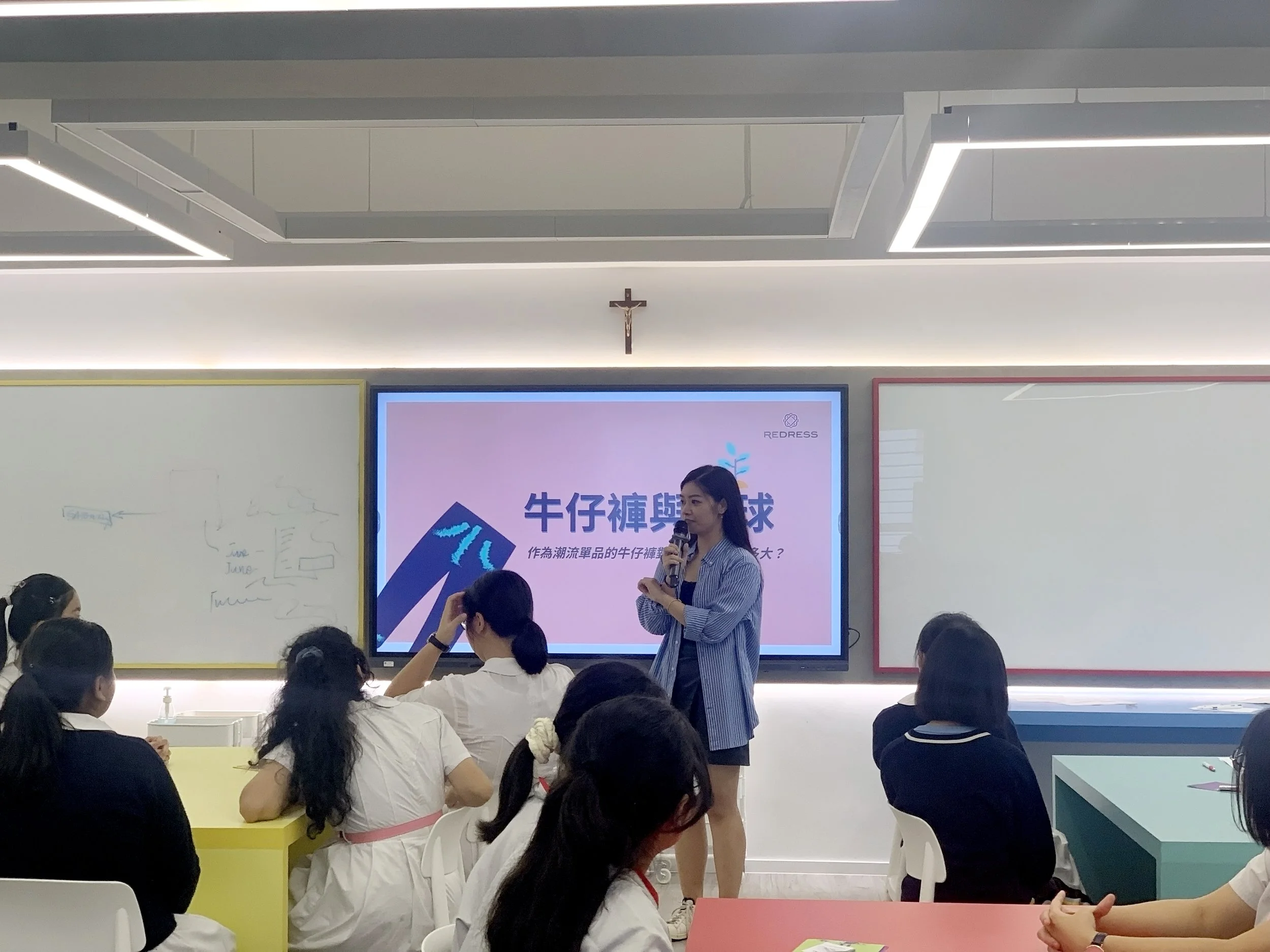A teacher giving a presentation to students in a classroom. The classroom has a large screen displaying text in Chinese and some whiteboards on either side. There is a crucifix mounted on the wall above the screen.