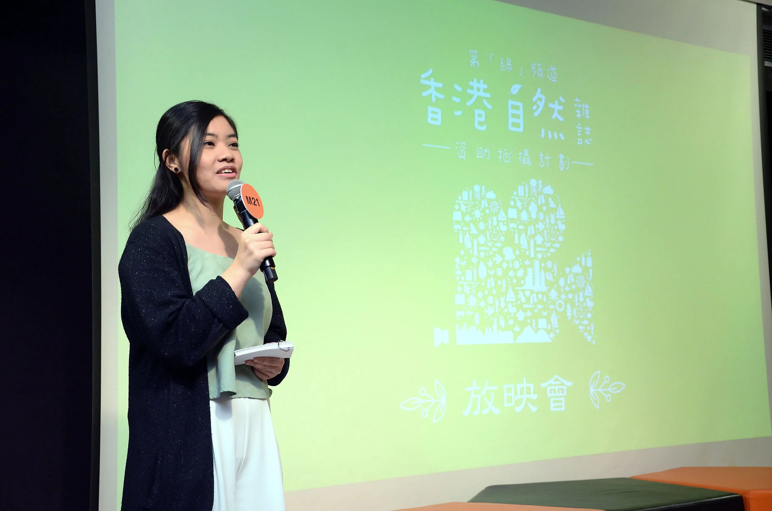 A young woman is standing and speaking into a microphone. She is holding some notes in her left hand. Behind her, there is a large screen displaying a graphic with Chinese characters and an illustration of a heart made up of various environmental-related icons on a light green background.