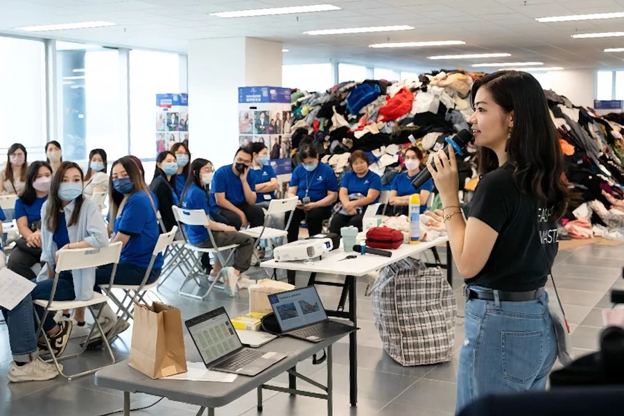A woman in a black shirt and jeans speaking into a microphone in front of a group of people in blue shirts, seated and wearing masks, in a room with large windows and a large pile of clothes in the background.