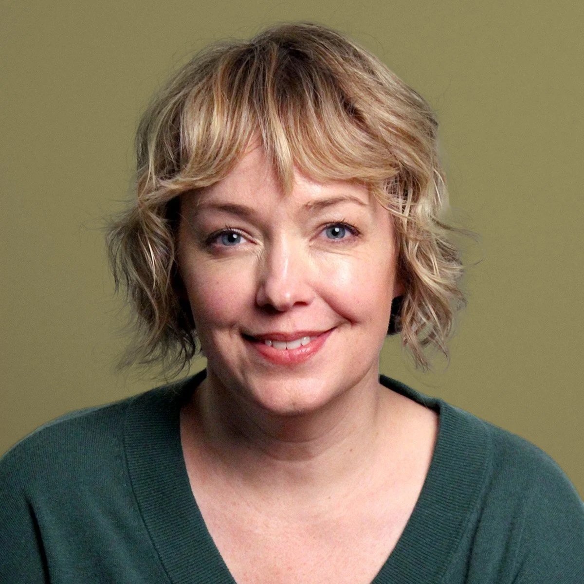 Nicole Kennelly, Professional Counselor Associate, Oregon, A woman with short, wavy blonde hair and blue eyes smiling at the camera, wearing a dark green top with a plain green background.