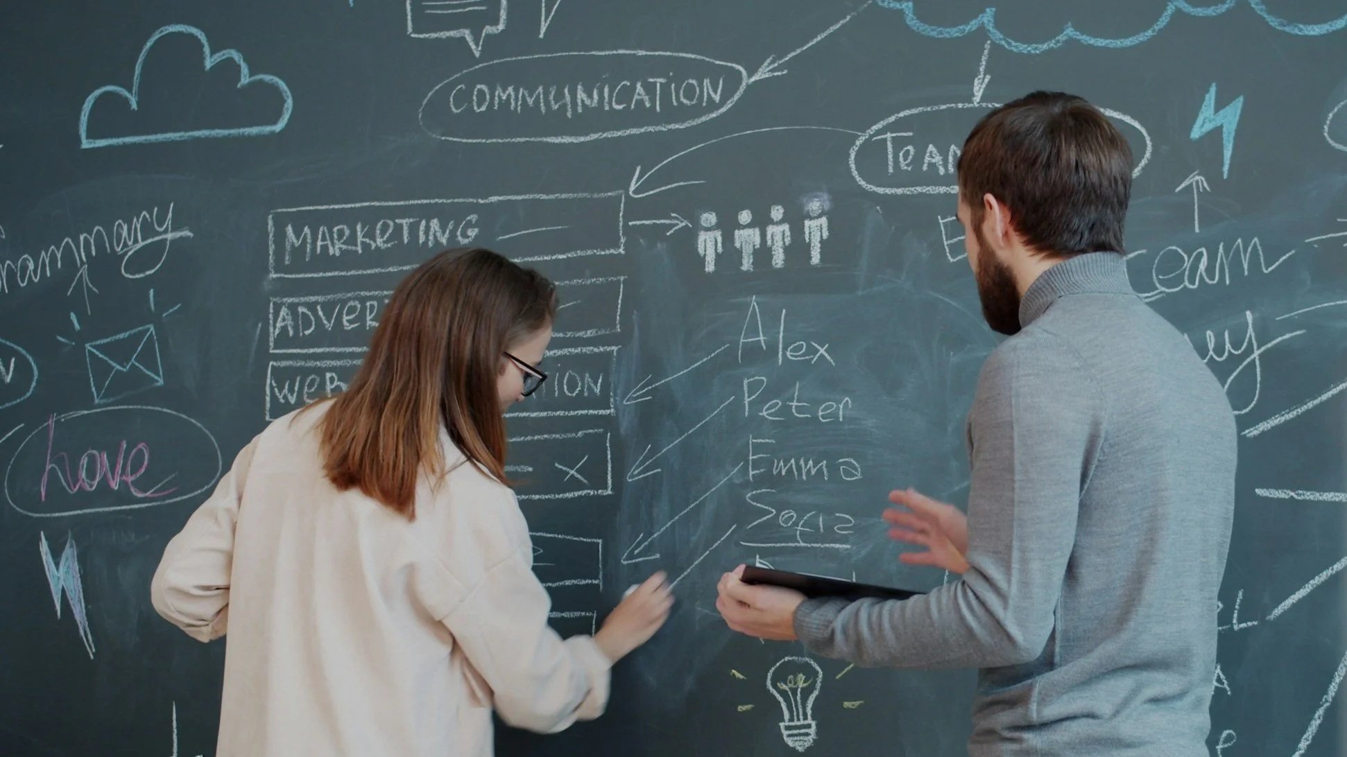 Two people, a woman and a man, discussing plans in front of a chalkboard filled with diagrams, text, and drawings related to marketing, communication, teamwork, and ideas.
