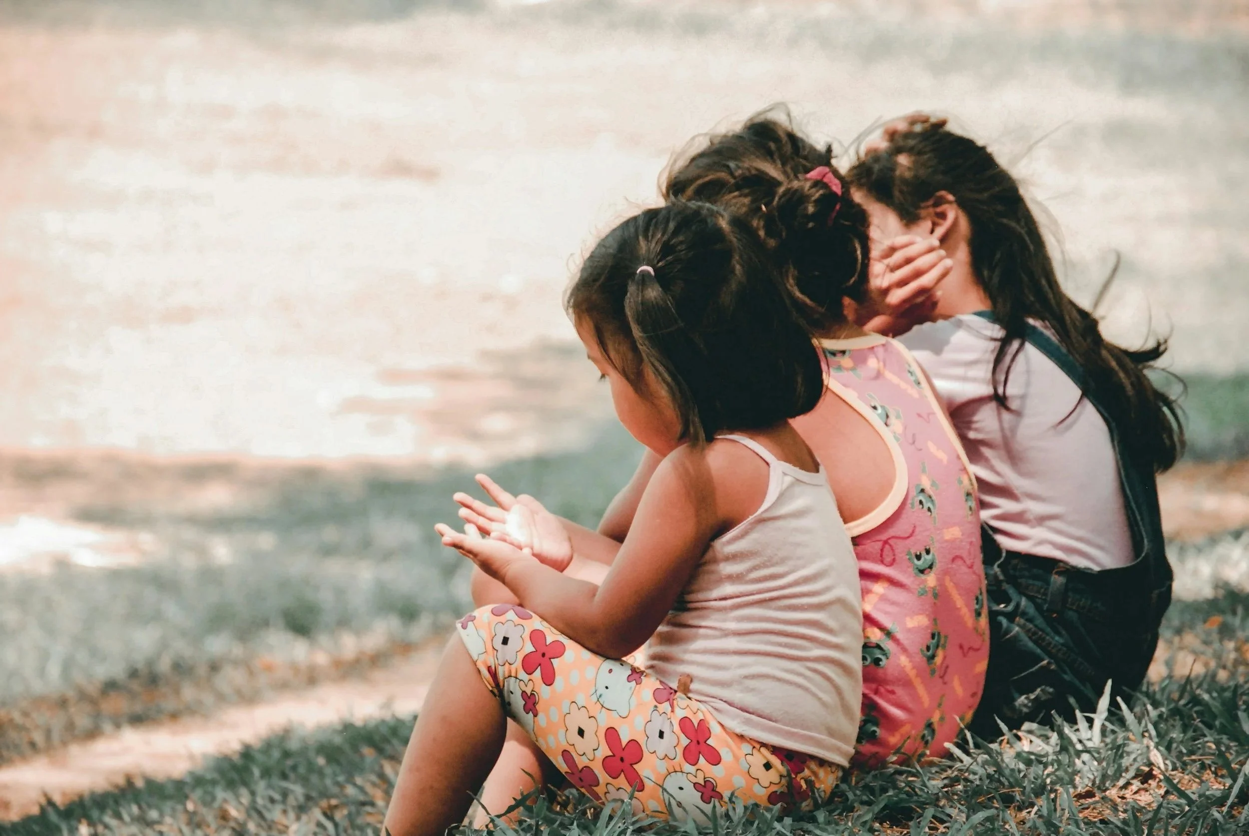 Three young girls sitting on the grass, two of them covering their faces and one looking at her hands.