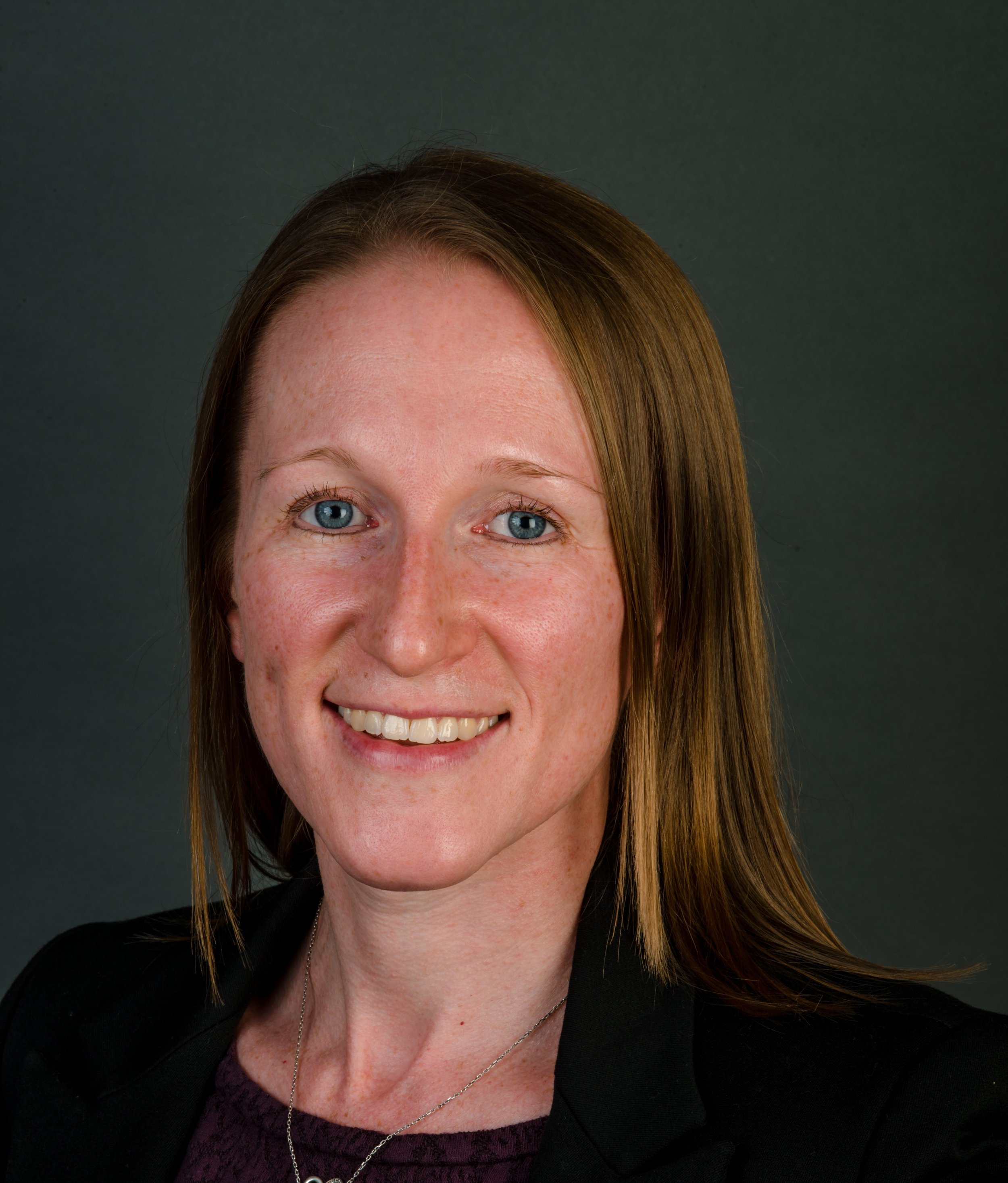 A woman with red hair, blue eyes, smiling, wearing a black blazer and a purple top, against a dark gray background.