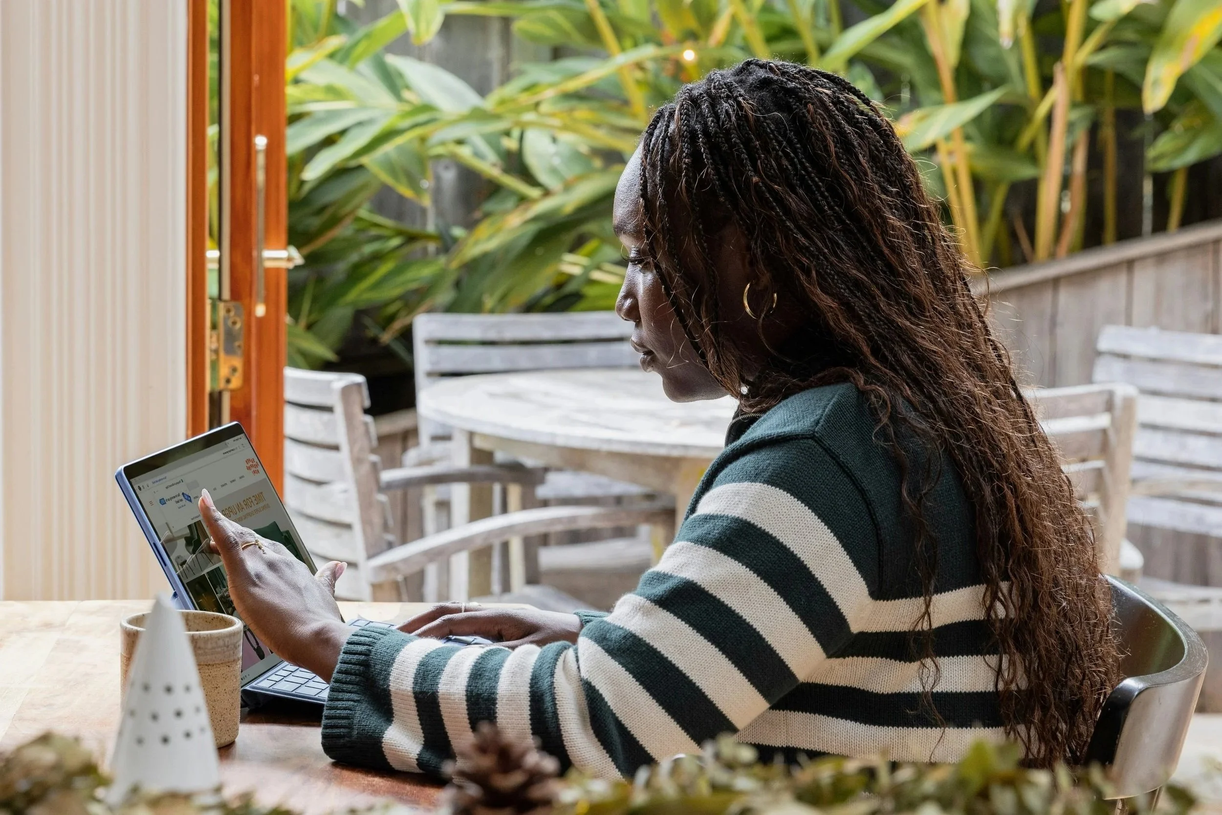 Woman with long curly hair sitting at a table, using a tablet in a garden or patio area with plants and outdoor furniture.