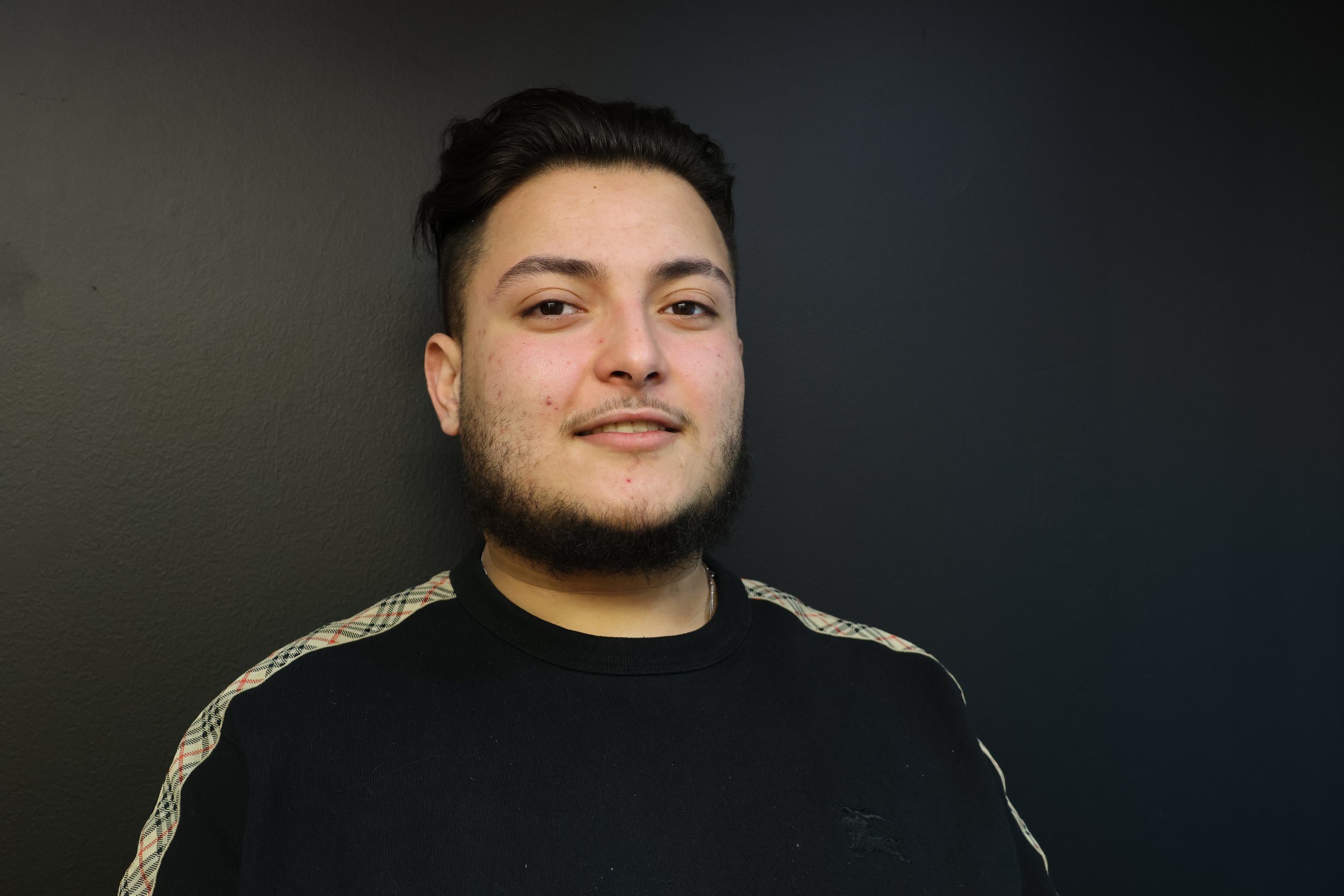 Portrait of a young man with dark hair and a beard, wearing a black shirt with plaid stripes on the shoulder, standing against a dark wall.