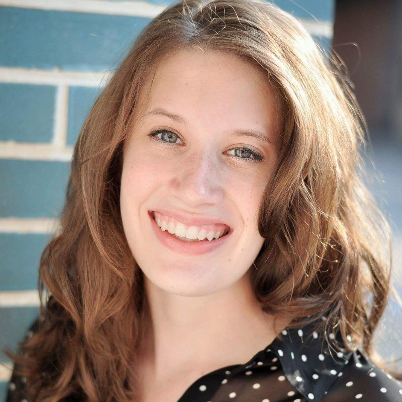 Close-up of a young woman with long, wavy auburn hair, smiling, wearing a black top with white polka dots, standing outdoors near a blue brick wall.