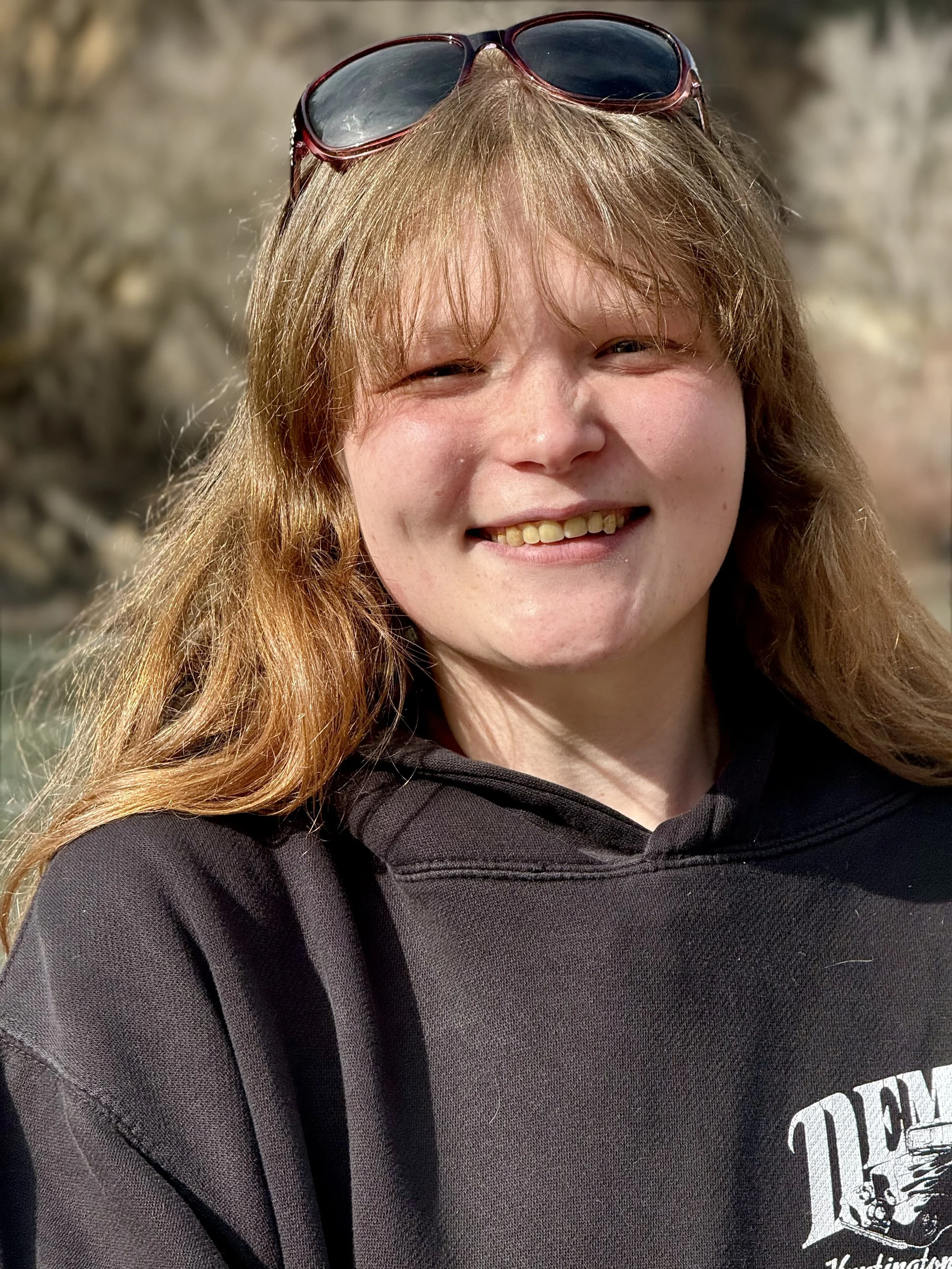 Close-up of a smiling woman with long, wavy, reddish-blonde hair wearing sunglasses and a black hoodie.