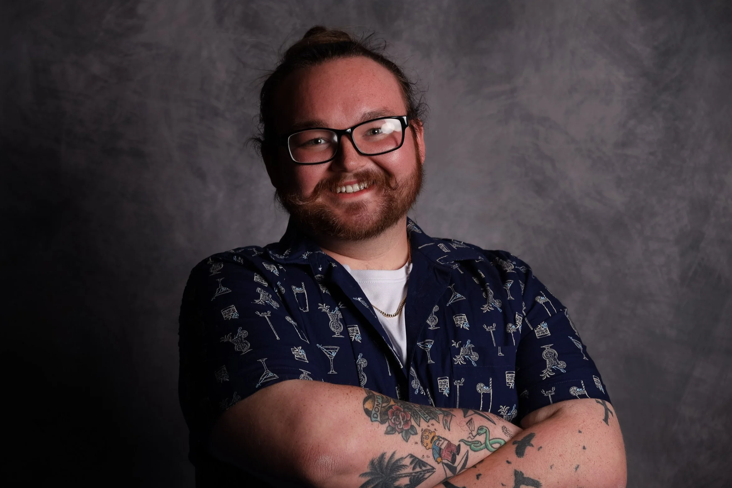 A man with glasses, a beard, and a mustache smiling, wearing a light pink shirt and a gold necklace with a Star of David pendant, against a dark background.
