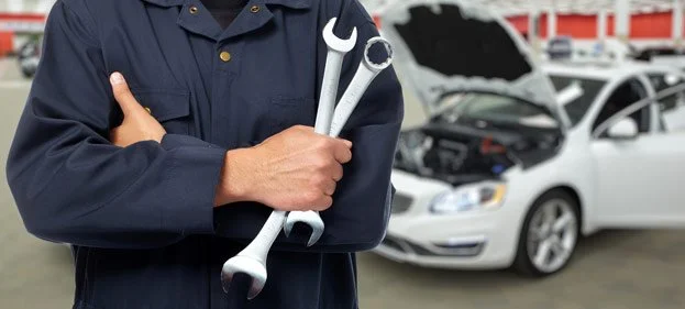 A mechanic wearing a navy blue uniform holding two wrenches in front of a white car with its hood open at a repair shop.