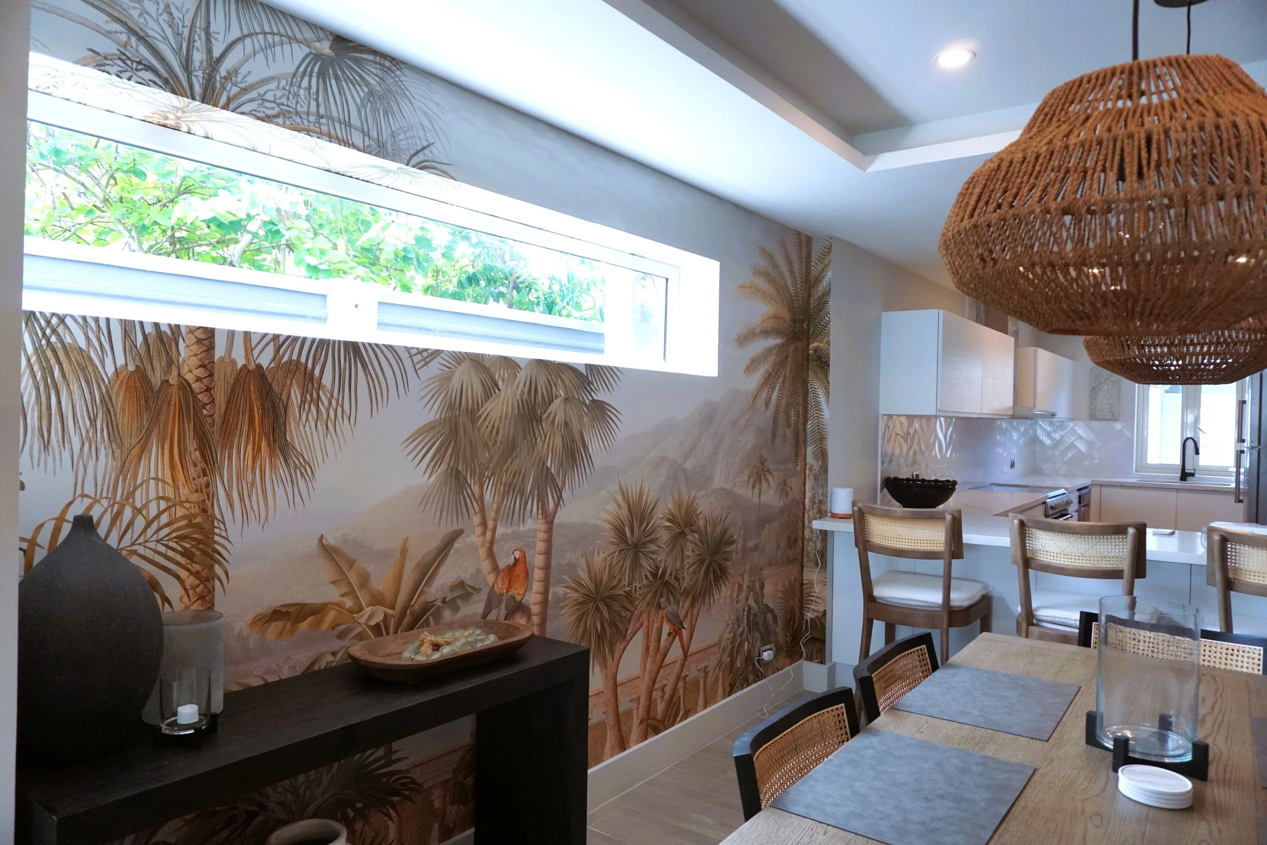 A dining area with a tropical jungle mural featuring palm trees on the wall, a large wicker pendant light, a wooden dining table with placemats, and chairs with woven backs. There's a black console table with decorative objects and an outdoor view through a horizontal window.