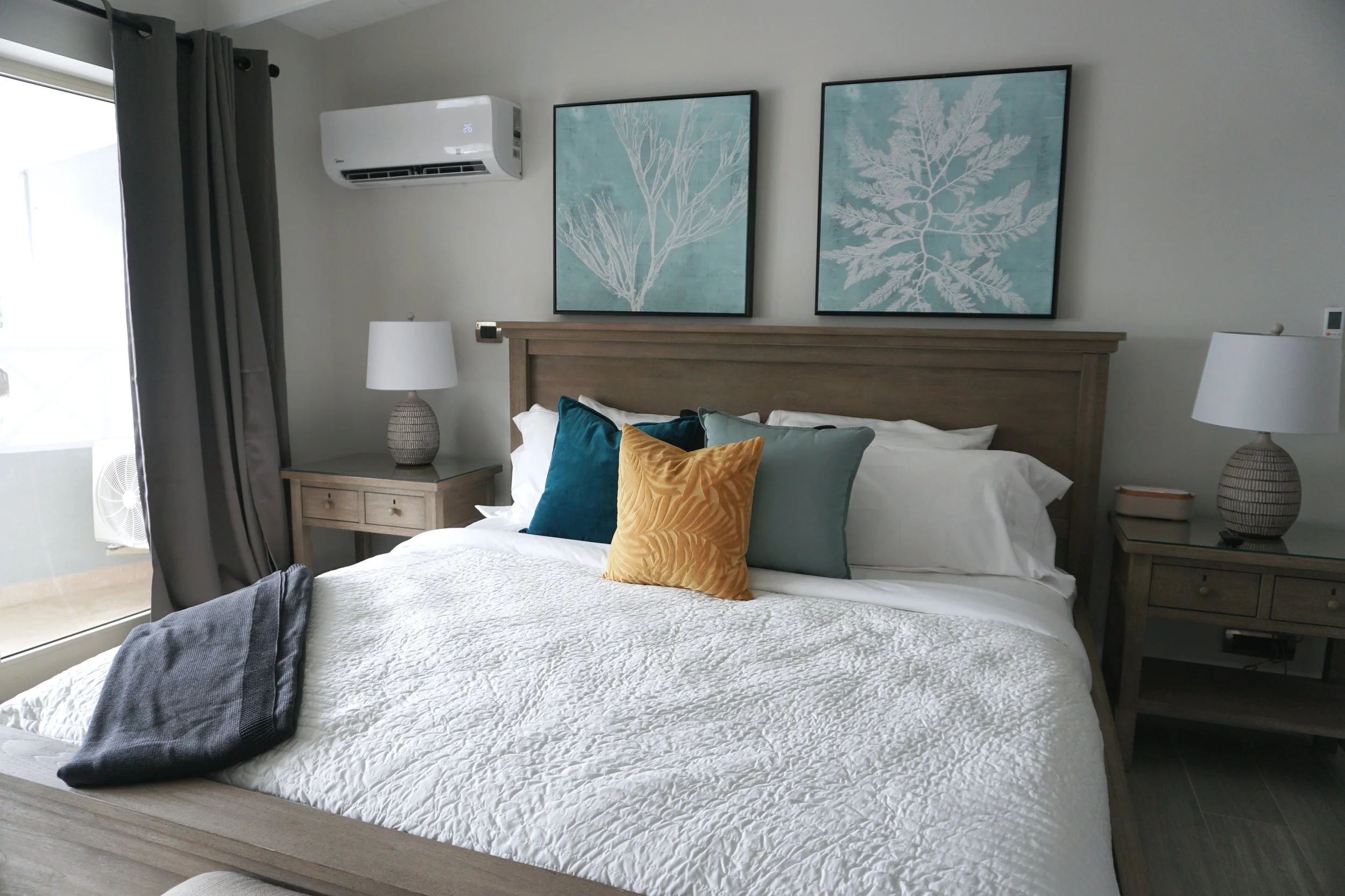 A neatly made bed with a gray wooden frame, decorated with white and dark-colored pillows. Two matching gray nightstands with table lamps, and two framed art prints of botanical illustrations hanging above the headboard in a bright, cozy bedroom.