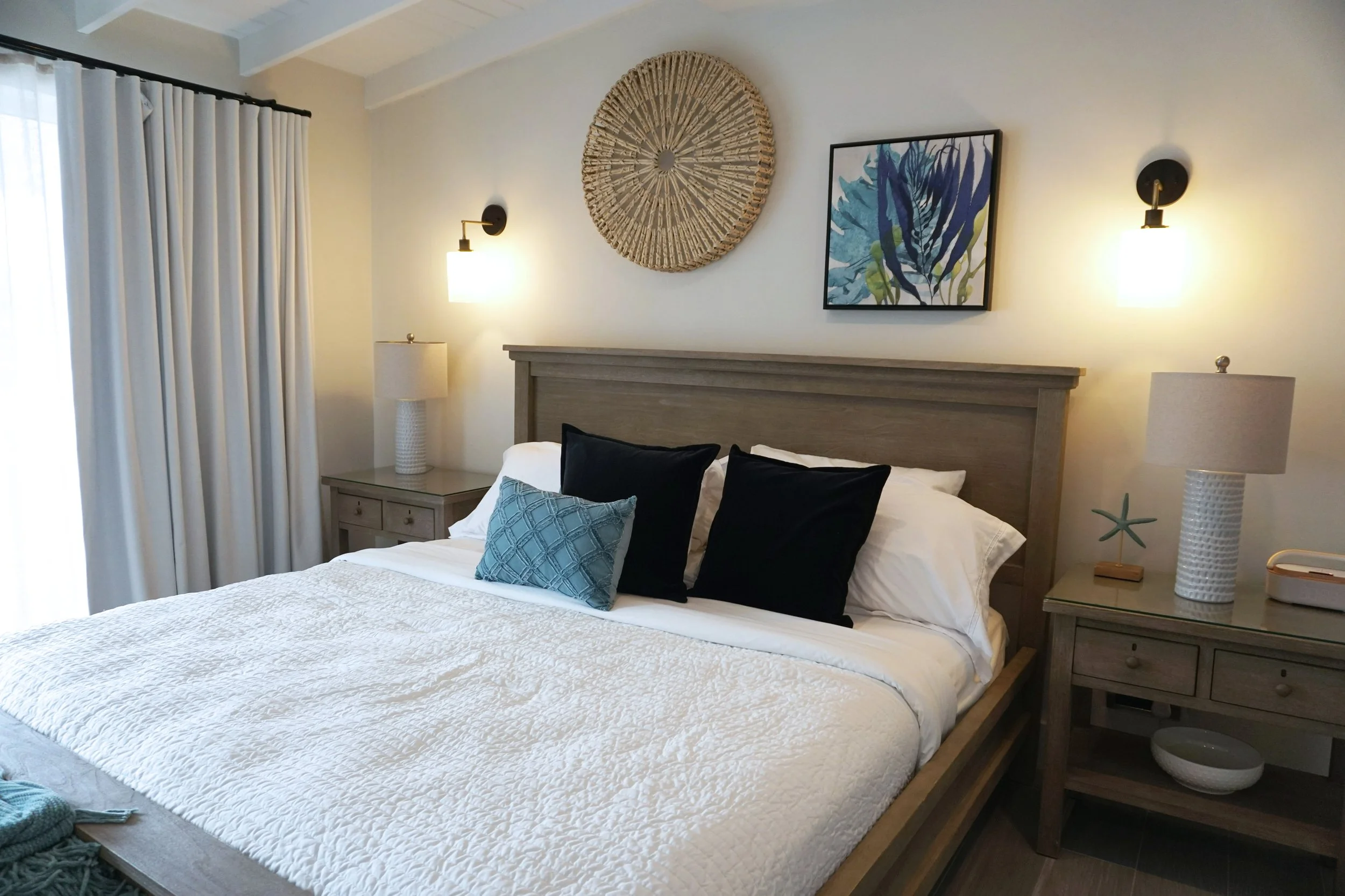 A bedroom with a bed that has white and dark pillows, a gray wooden headboard, and a light quilt. There are two black nightstands with lamps, a black pouf at the foot of the bed, and artwork and wall decor above the headboard. A sliding glass door with gray curtains leads to a balcony.