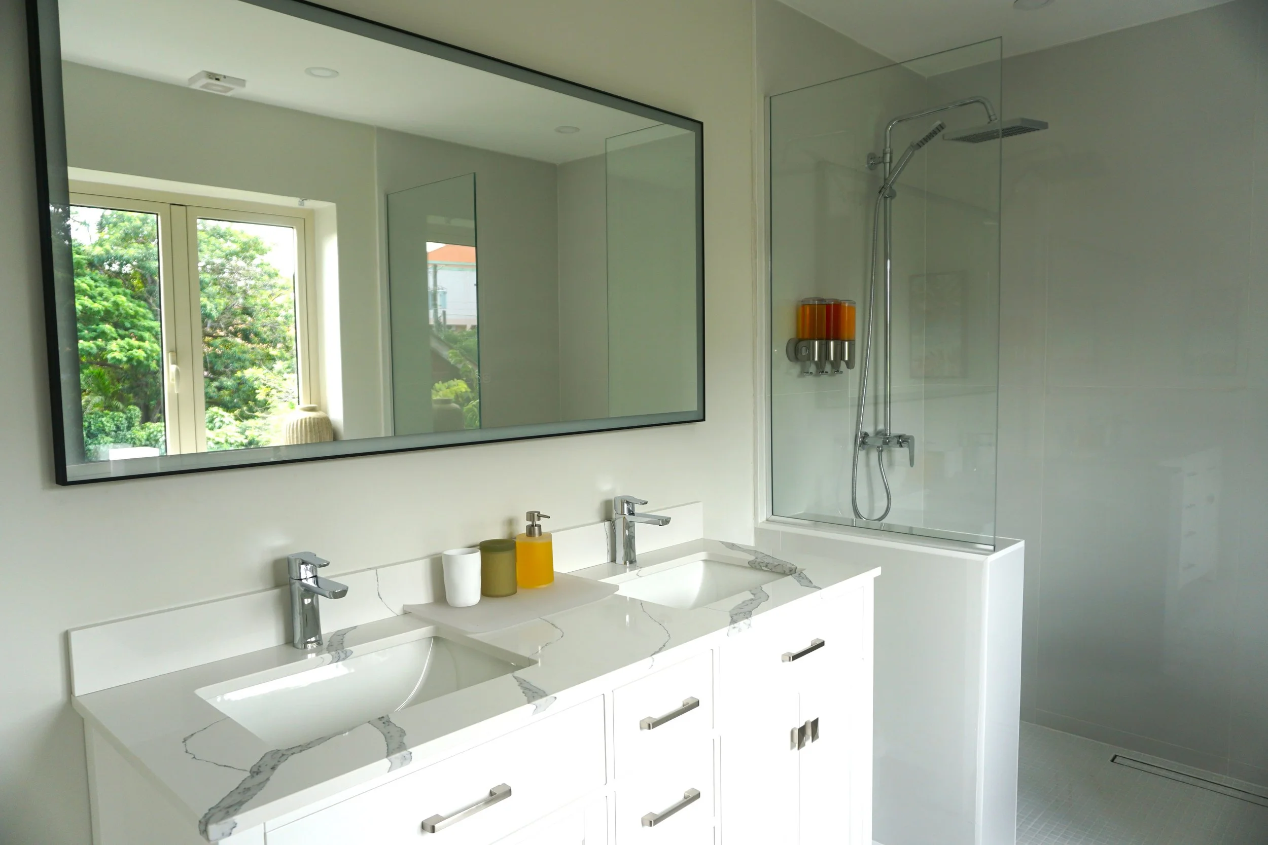 A clean, modern bathroom with a double vanity, large mirror, glass shower enclosure, and a toilet with a white tank and seat. The floor and walls are light-colored, and there are recessed ceiling lights.