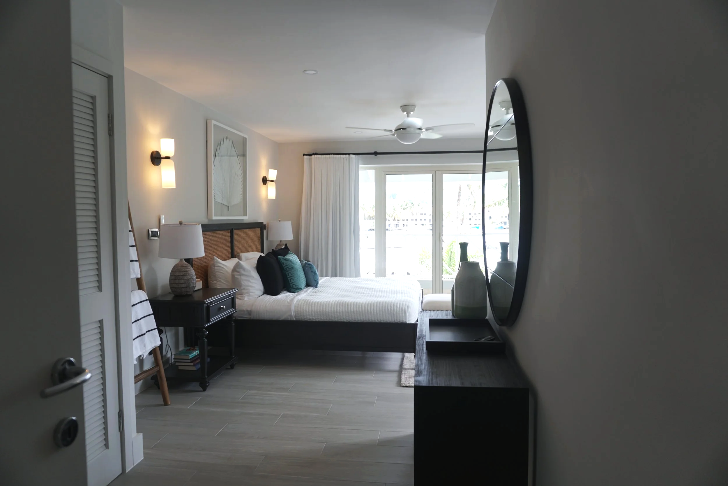 A modern bedroom with a large bed, white bedding, and teal and black pillows. There are two wall-mounted lamps, a bedside table with a lamp, and a decorative piece above the bed. A sliding glass door with white curtains leads to a balcony with outdoor scenery. On the right side, a black console table with three decorative vases and a curved black mirror reflect part of the room.