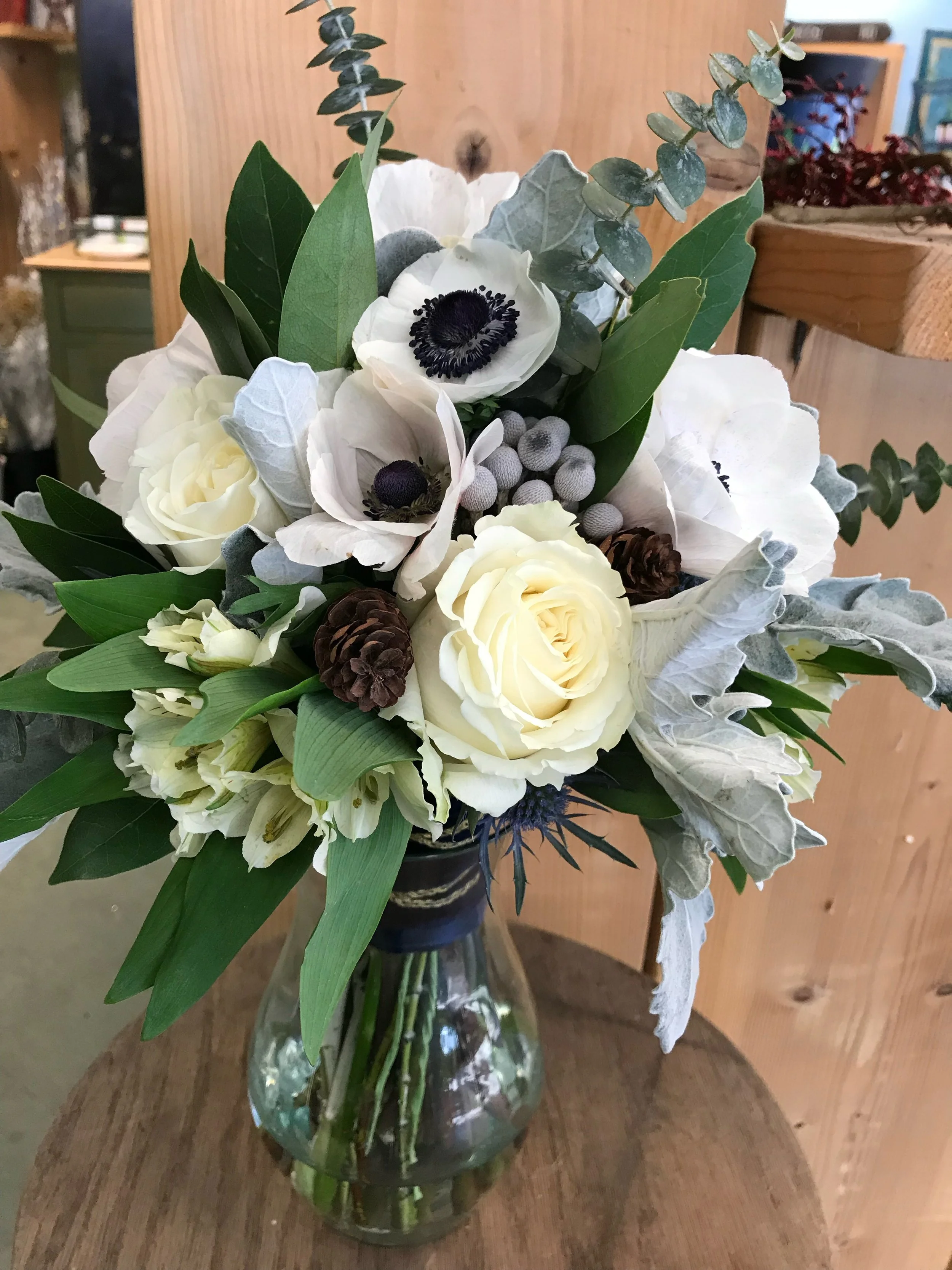 A floral arrangement with white roses, anemones, eucalyptus, and pinecones in a glass vase on a wooden table.