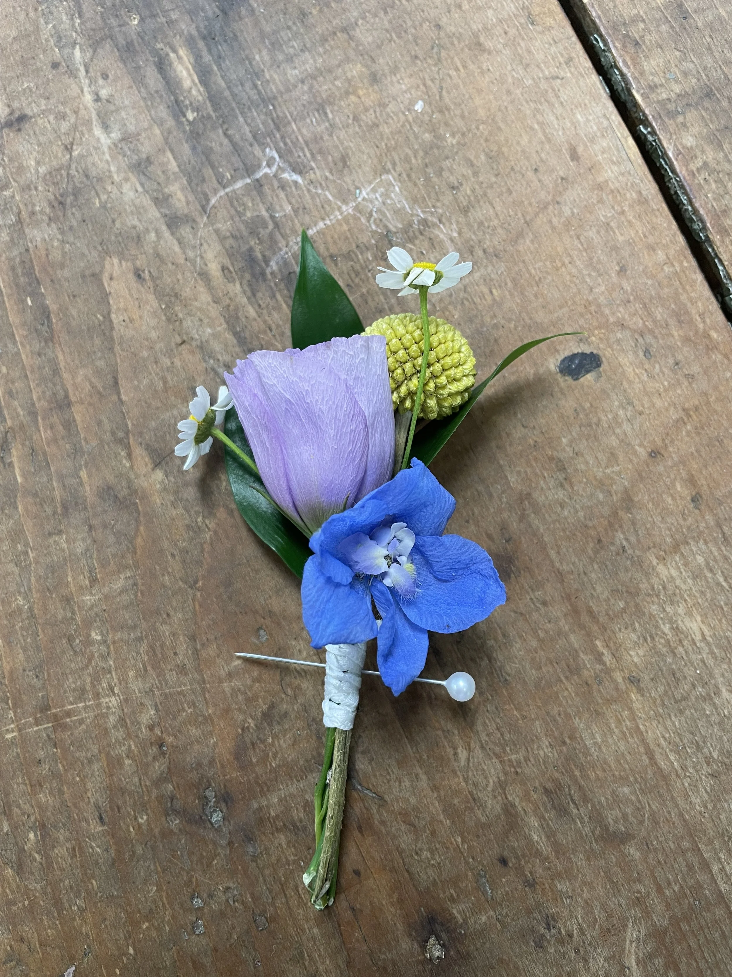A boutonniere with a purple tulip, blue iris, small white daisies, and yellow clustered flowers, pinned with a white pearl pin on a wooden surface.