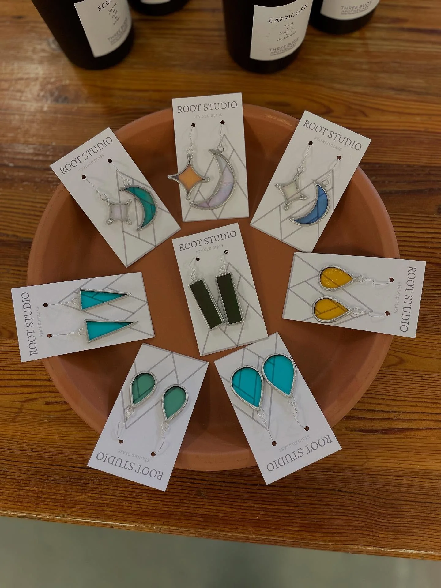 Collection of colorful glass earrings from Root Studio arranged on a round wooden display with bottles in the background.