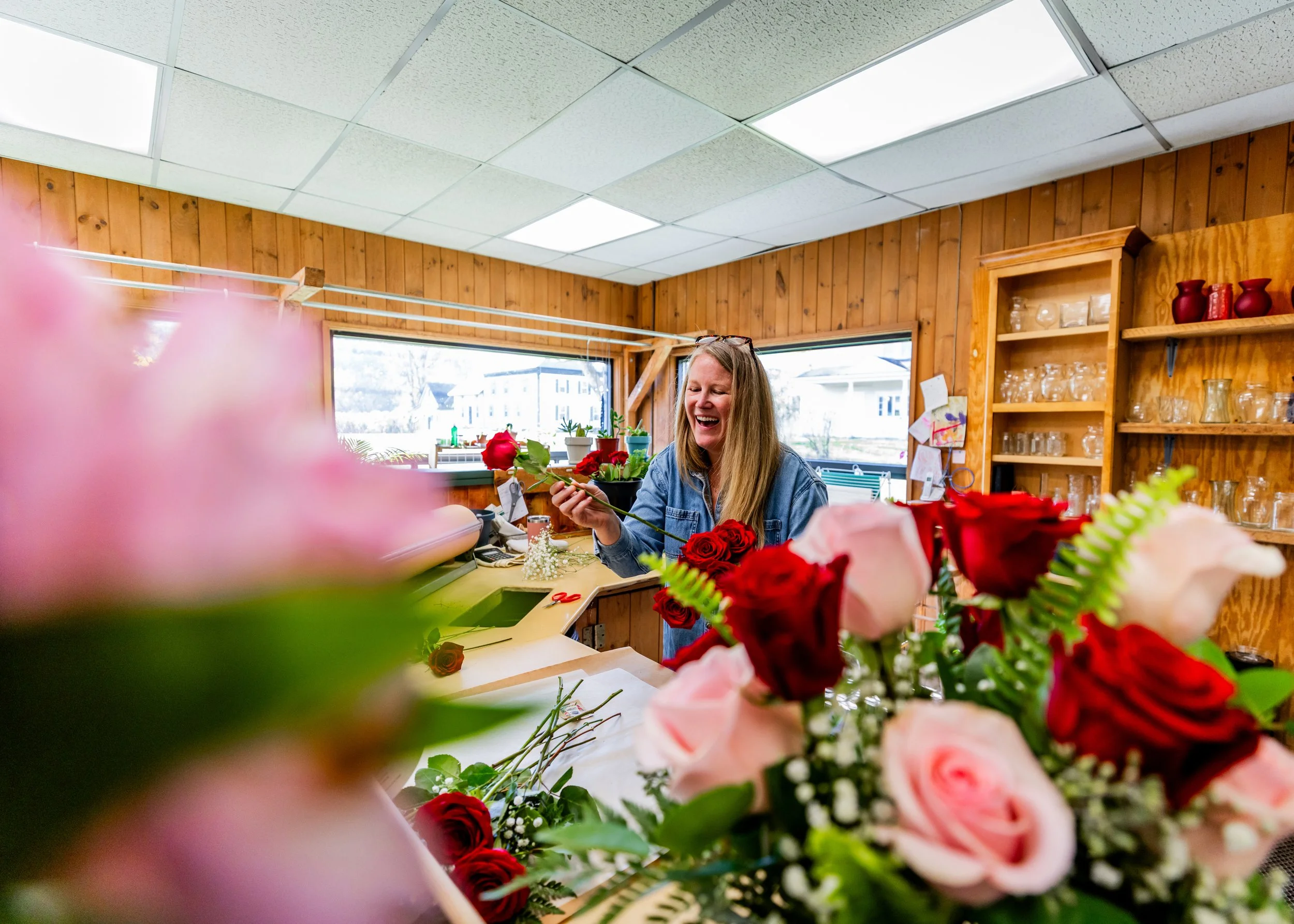 Woman smiling and working with a bunch of red and pink roses in a flower shop with wooden walls and shelves filled with glassware.